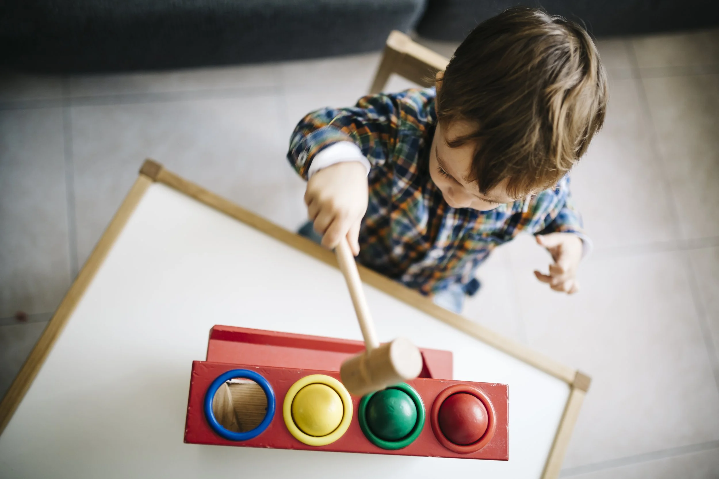 Child in plaid shirt playing with a toy hammer and colored peg board.