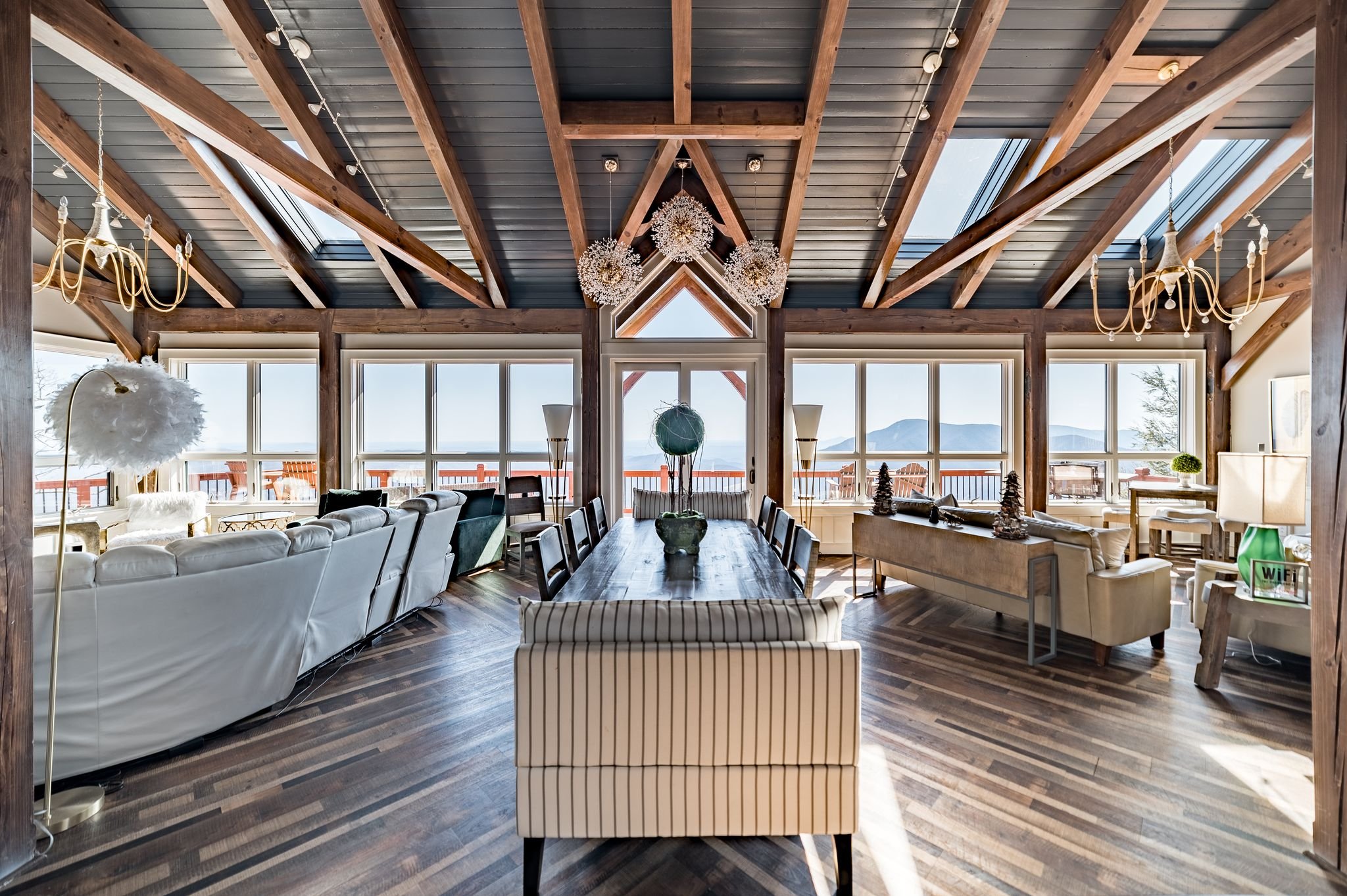 Spacious modern living room with large windows, wooden beams, and a vaulted ceiling. Features a dining table, white sofas, pendant lights, and a view of mountains.