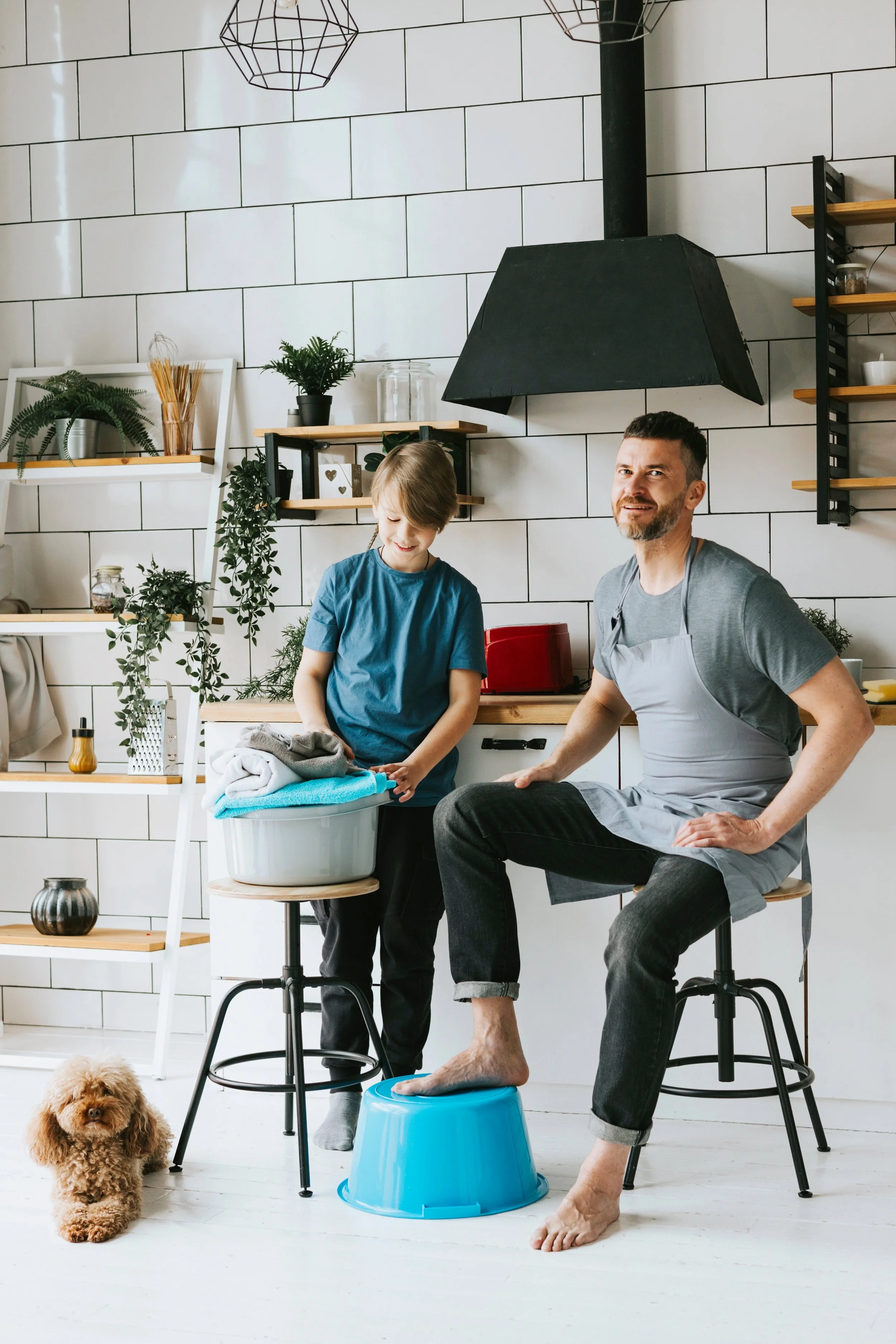A man with a beard, wearing a gray apron, sitting on a stool with his bare foot on a blue bucket. A child in a blue shirt stands nearby, smiling, with folded towels in a laundry basket. A small brown dog sits on the floor in a modern kitchen with plants and white tiles.