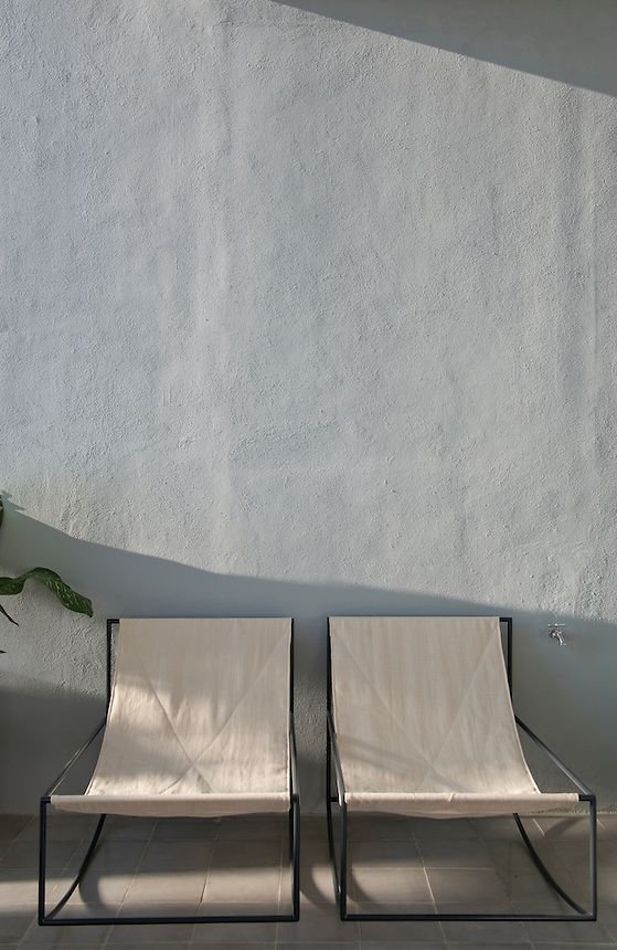 Two modern beige chairs against a gray wall, with a small plant on the side, casting soft shadows on a tiled floor.
