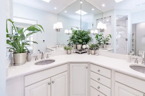 Modern white bathroom with dual sinks, large mirrors, and multiple potted plants. The room has a glass shower door and recessed lighting.