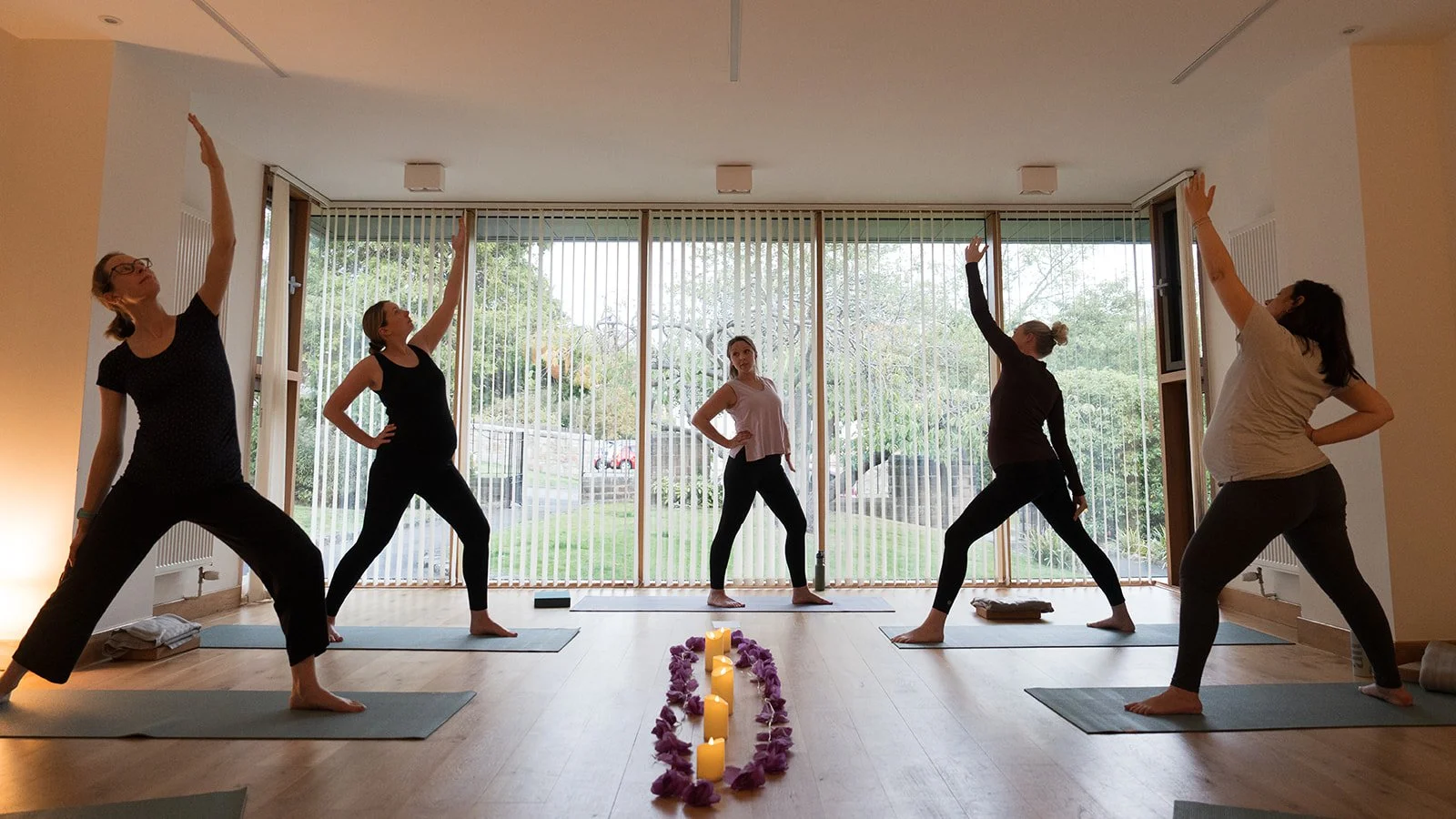 Four pregnant women and their yoga teacher Louise Burchell stand in reverse warrior. There are LED candles and purple flowers in the centre of the room.