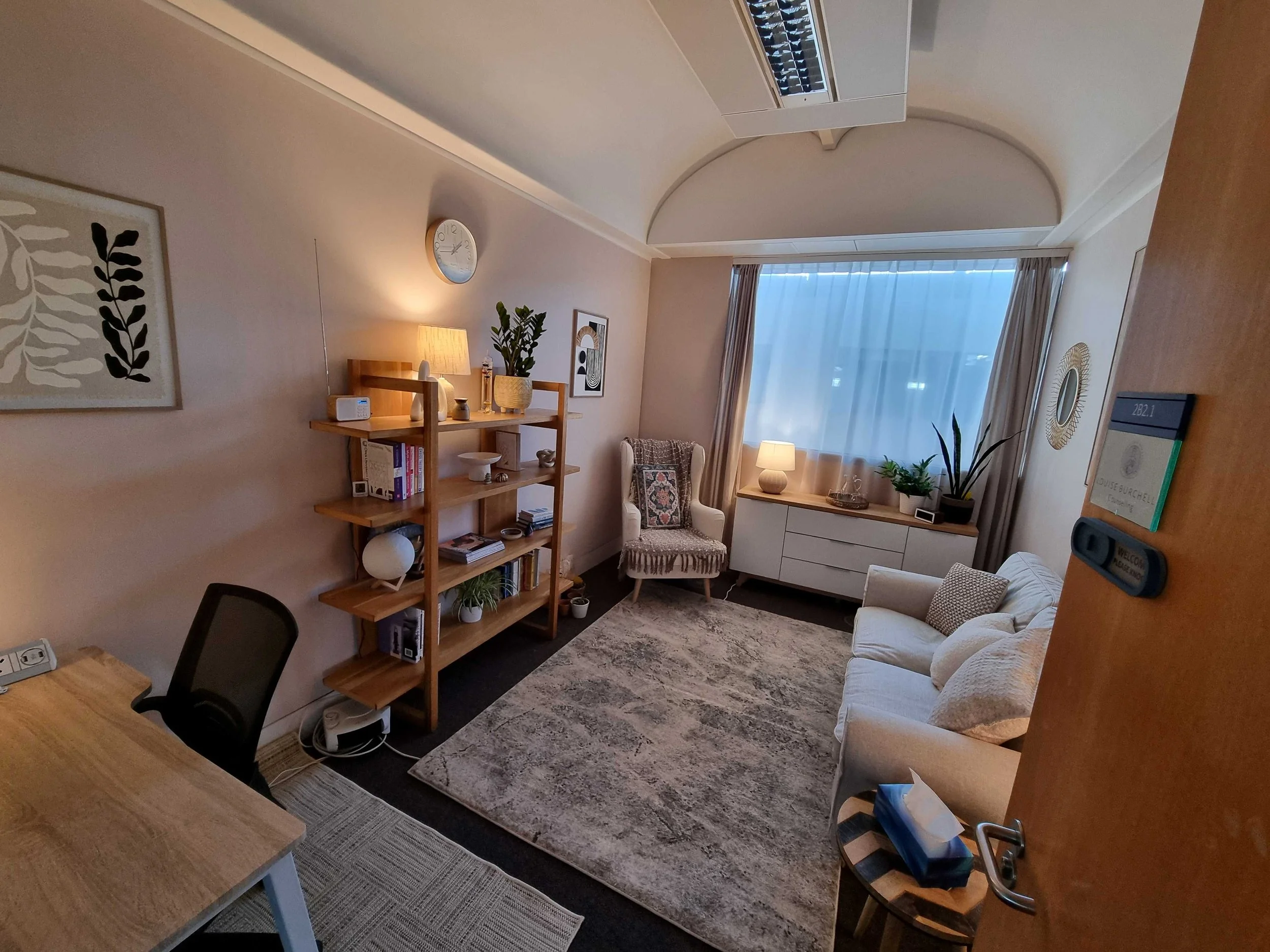 A desk and chair at the bottom left, with large bookcase showcasing books and ornaments, an armchair, sideboard and comy sofa. cosy rug