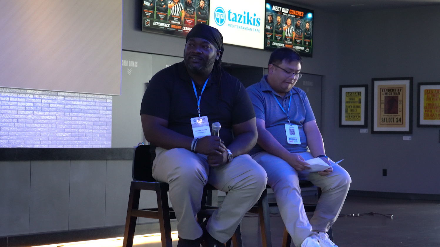 Two men sit on stools, one holds mic as they present