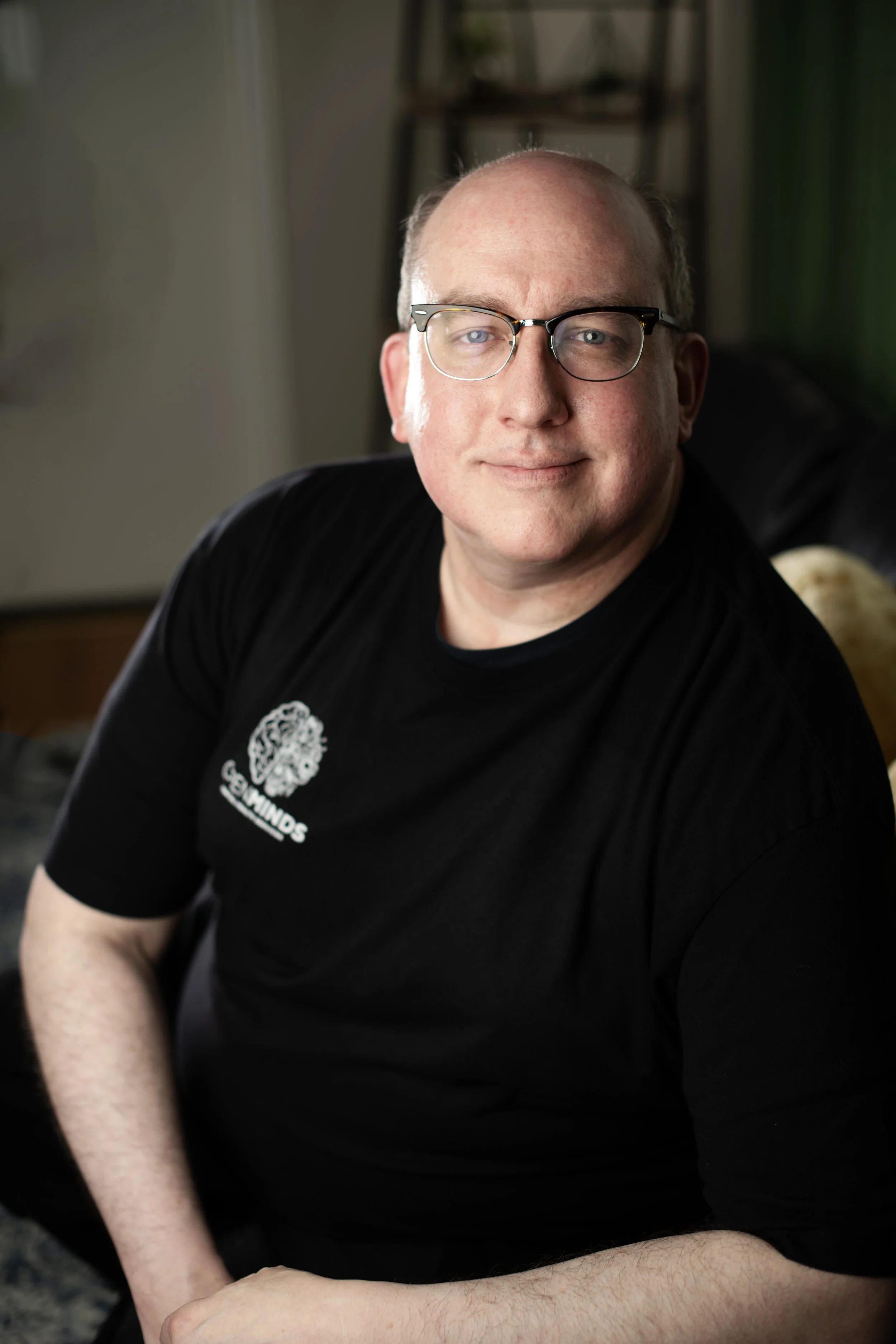 A man with glasses and a black t-shirt sitting on a couch, looking at the camera with a slight smile.