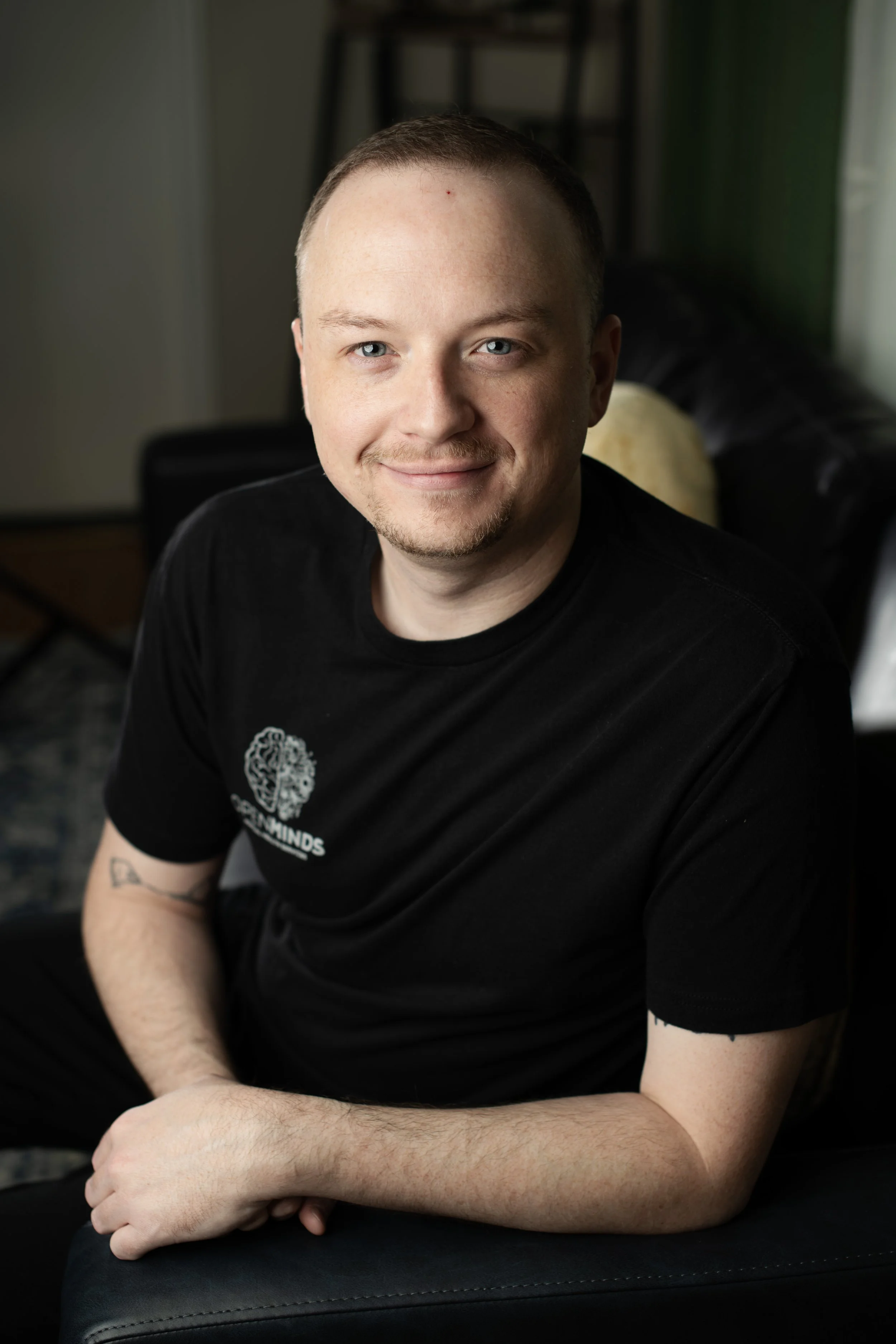 A young man with short hair and a goatee, wearing a black T-shirt, sitting on a chair in a room with a bookshelf and green wall in the background, smiling at the camera.