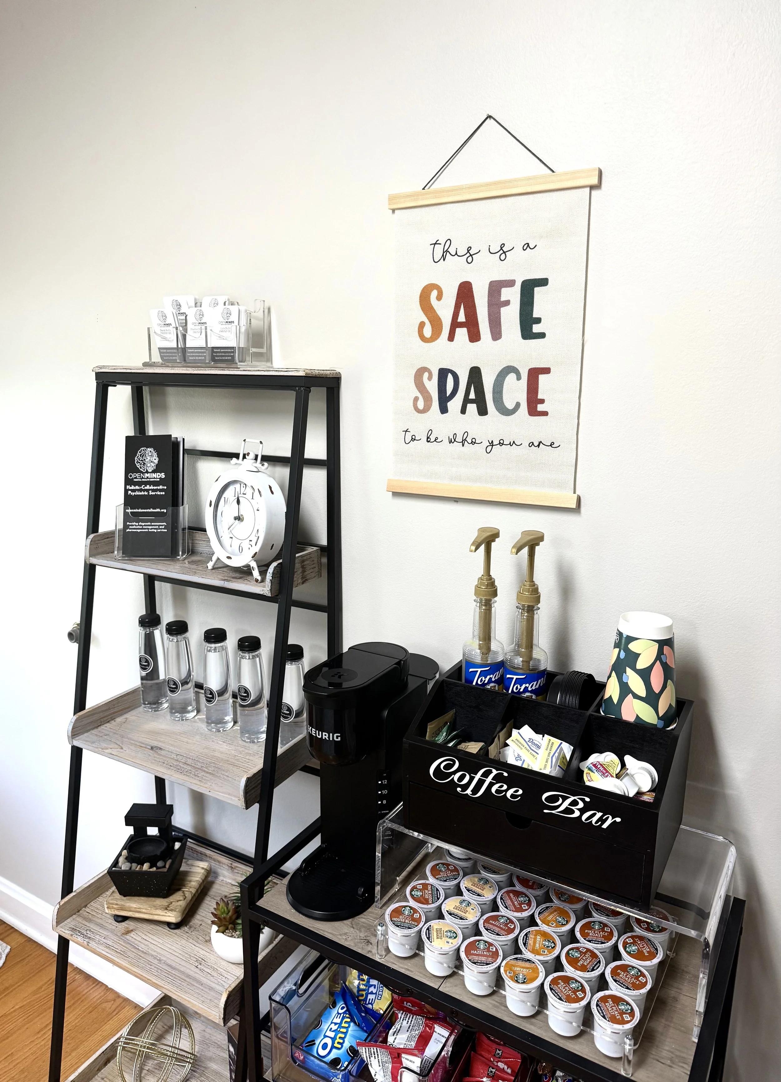 Coffee station with a Keurig machine, coffee pods, lids, and stir sticks, on a multi-shelf rack with glasses, a clock, and promotional materials; a sign on the wall reads 'this is a SAFE SPACE to be who you are.'