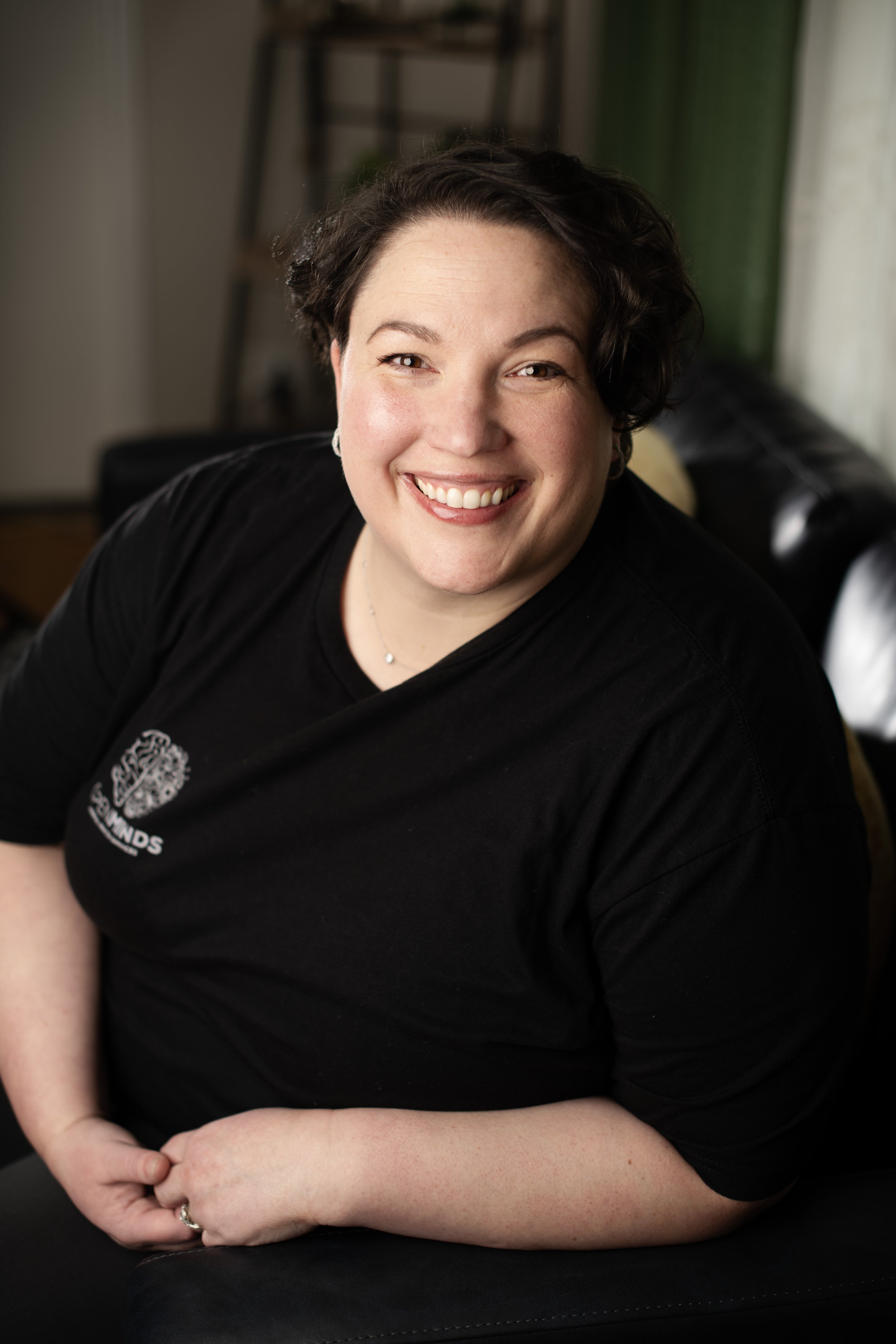 A woman with short dark hair smiling and wearing a black t-shirt, sitting in a cozy room with a ladder and curtains in the background.