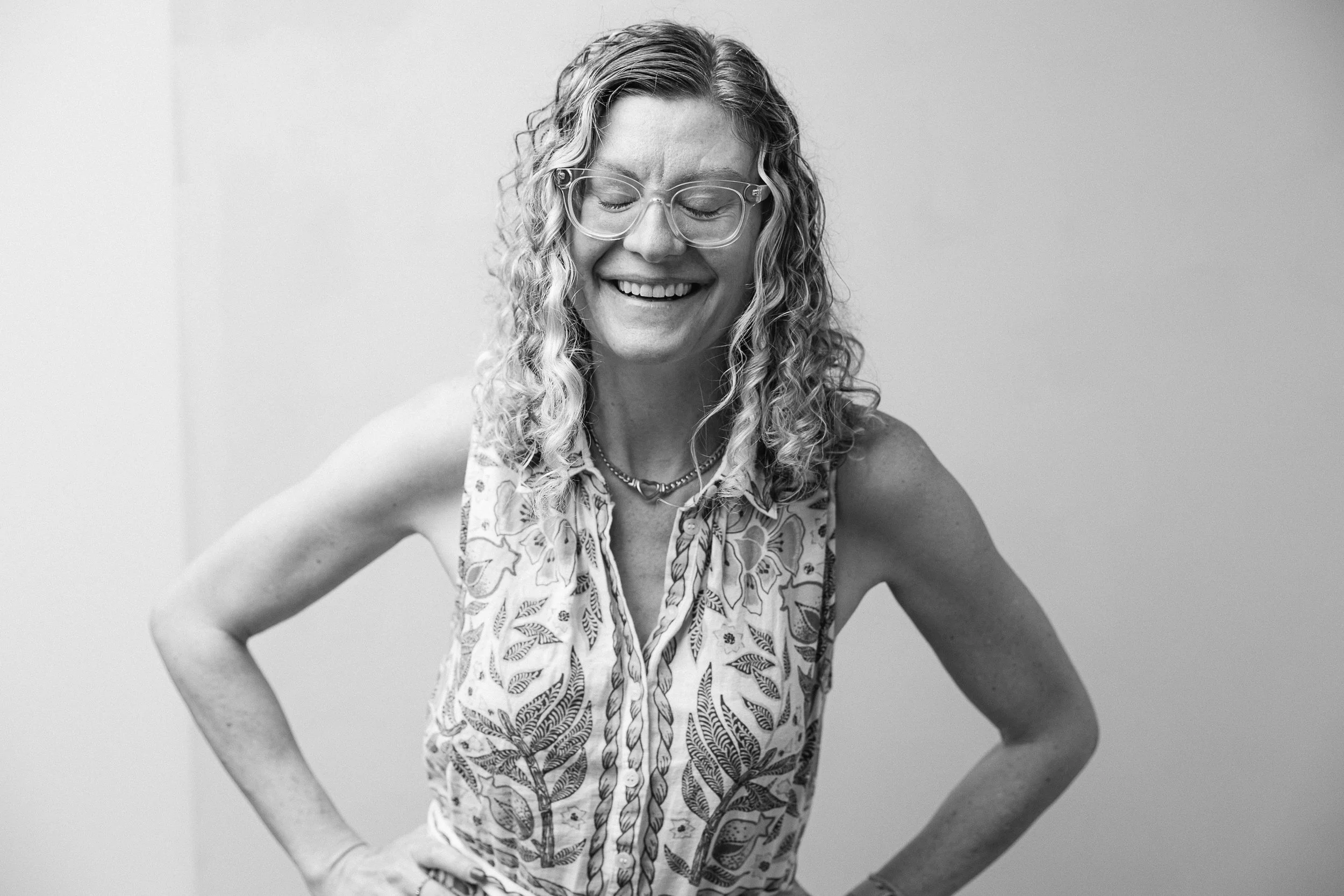 A woman with curly hair, glasses, and a patterned sleeveless top smiling with her eyes closed, standing against a plain wall. Photo by Liza Hippler.