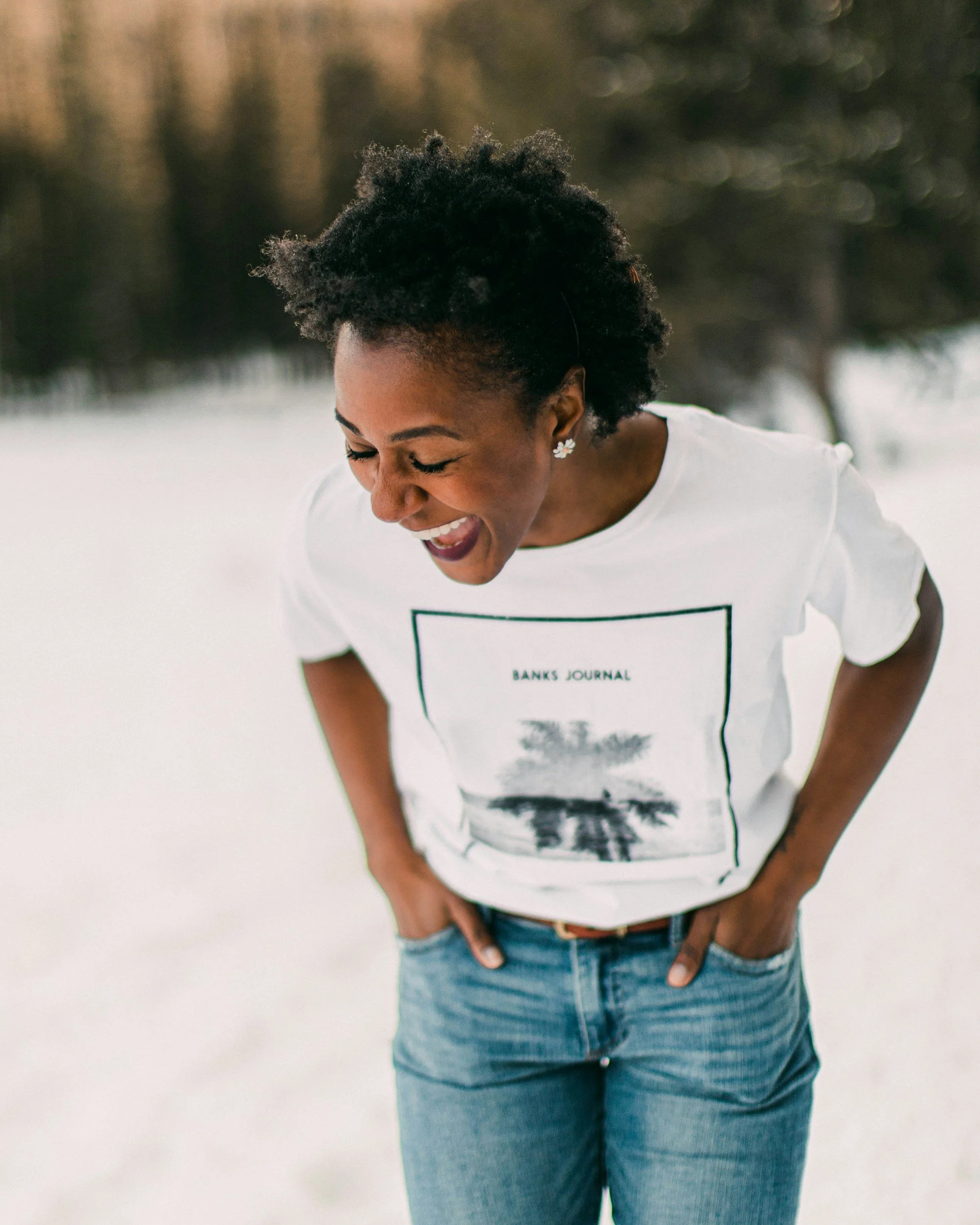 A woman laughing outdoors, wearing a white t-shirt with a graphic print and light blue jeans, with her hands in her pockets, short curly hair, and earrings, on a blurry background.