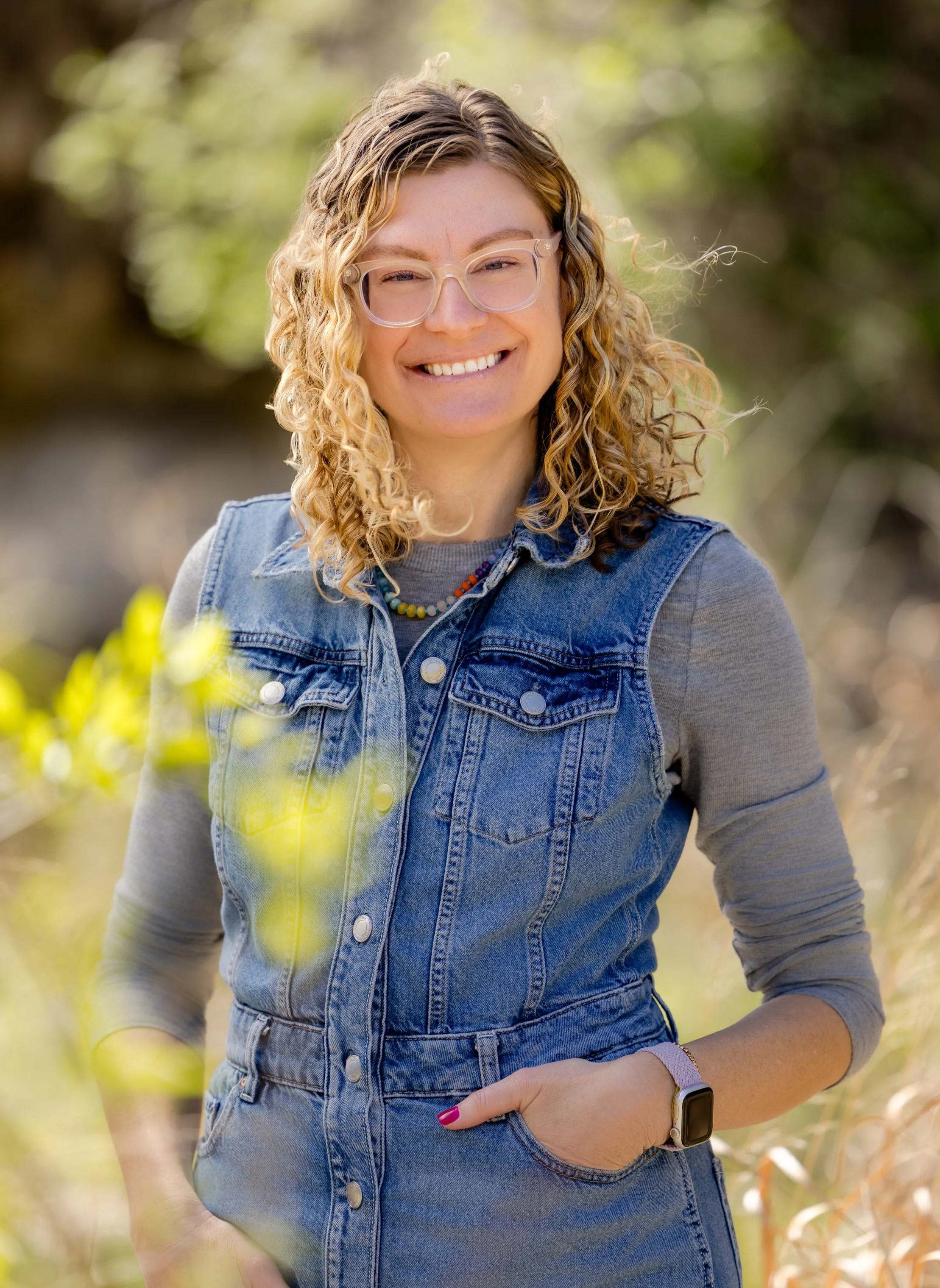 A woman with curly blonde hair and glasses smiling outdoors in a park, wearing a denim vest over a gray shirt, a colorful beaded necklace, a pink watch, and a smartwatch on her left wrist.