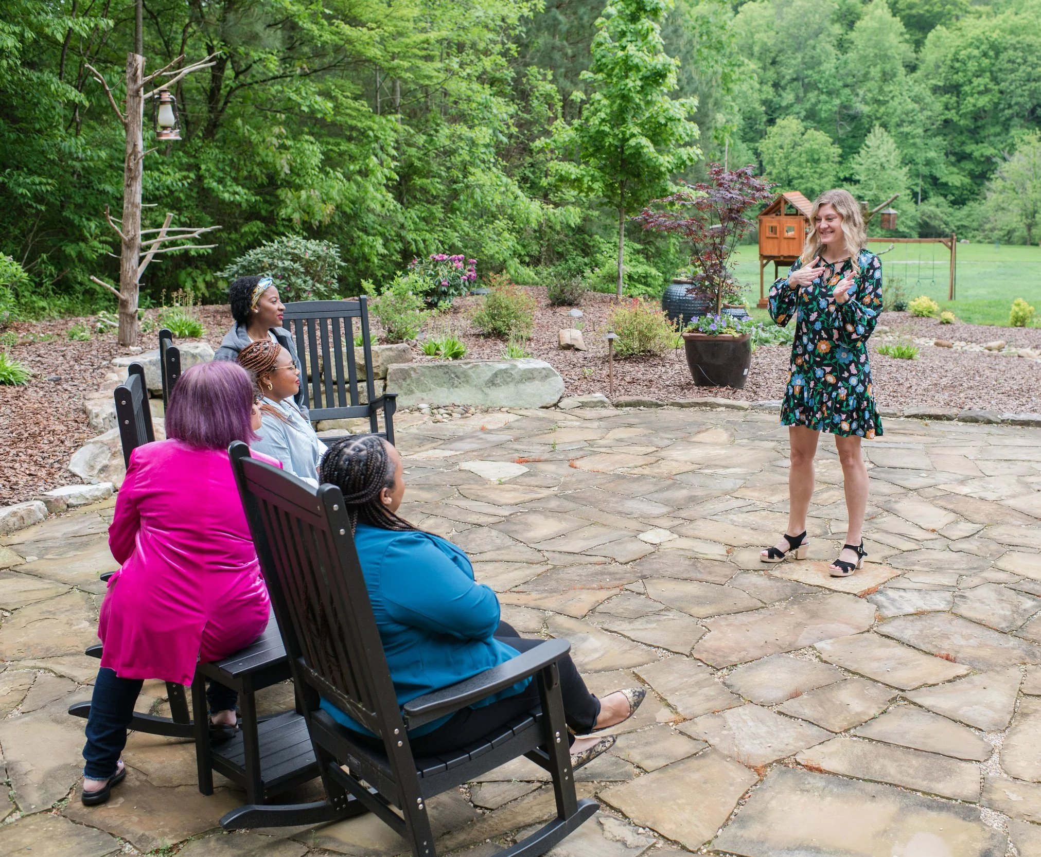 A woman with wavy blonde hair wearing a dress with a floral pattern is standing outdoors near a garden with trees and shrubs, smiling and gesturing with her hands while speaking to a group of seated women in colorful clothing.