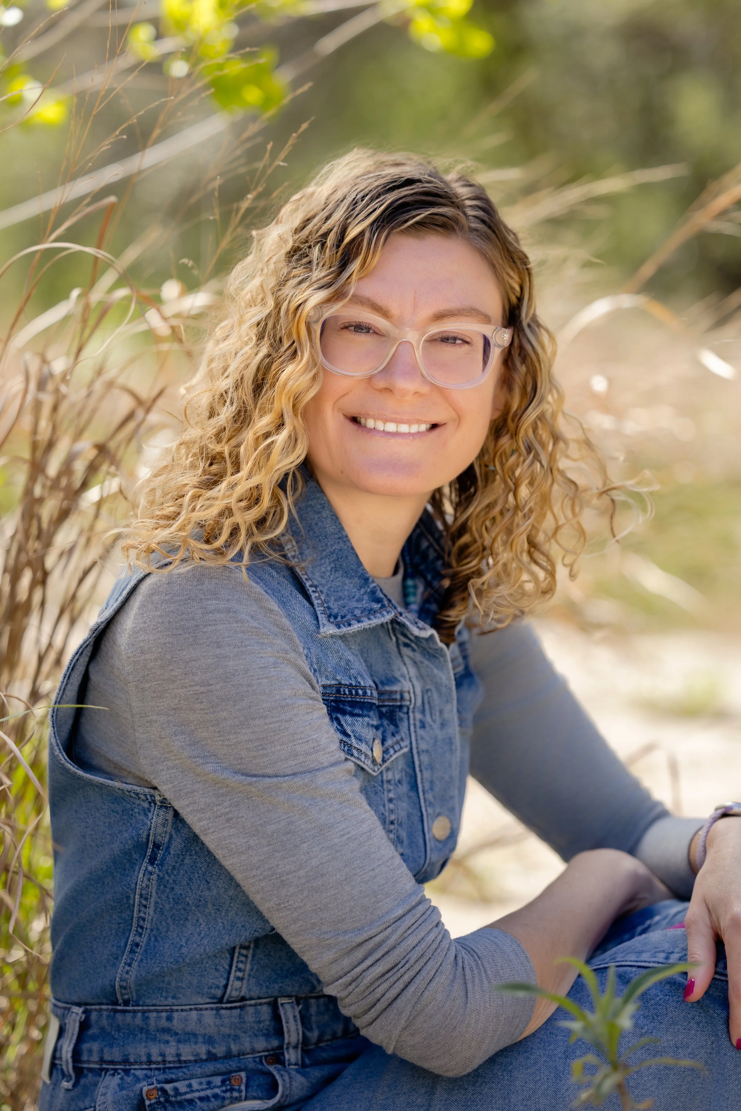 A woman with curly blonde hair and glasses sitting outdoors in a natural setting, smiling at the camera.