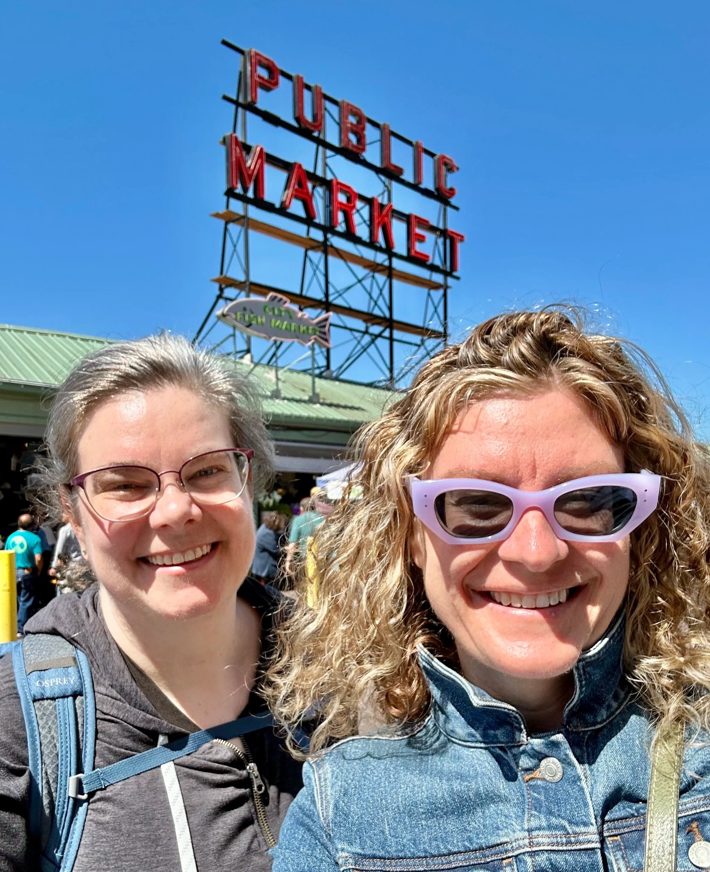 Two sisters in front of Seattle's Pike Place Market on a sunny day with a bright blue sky.