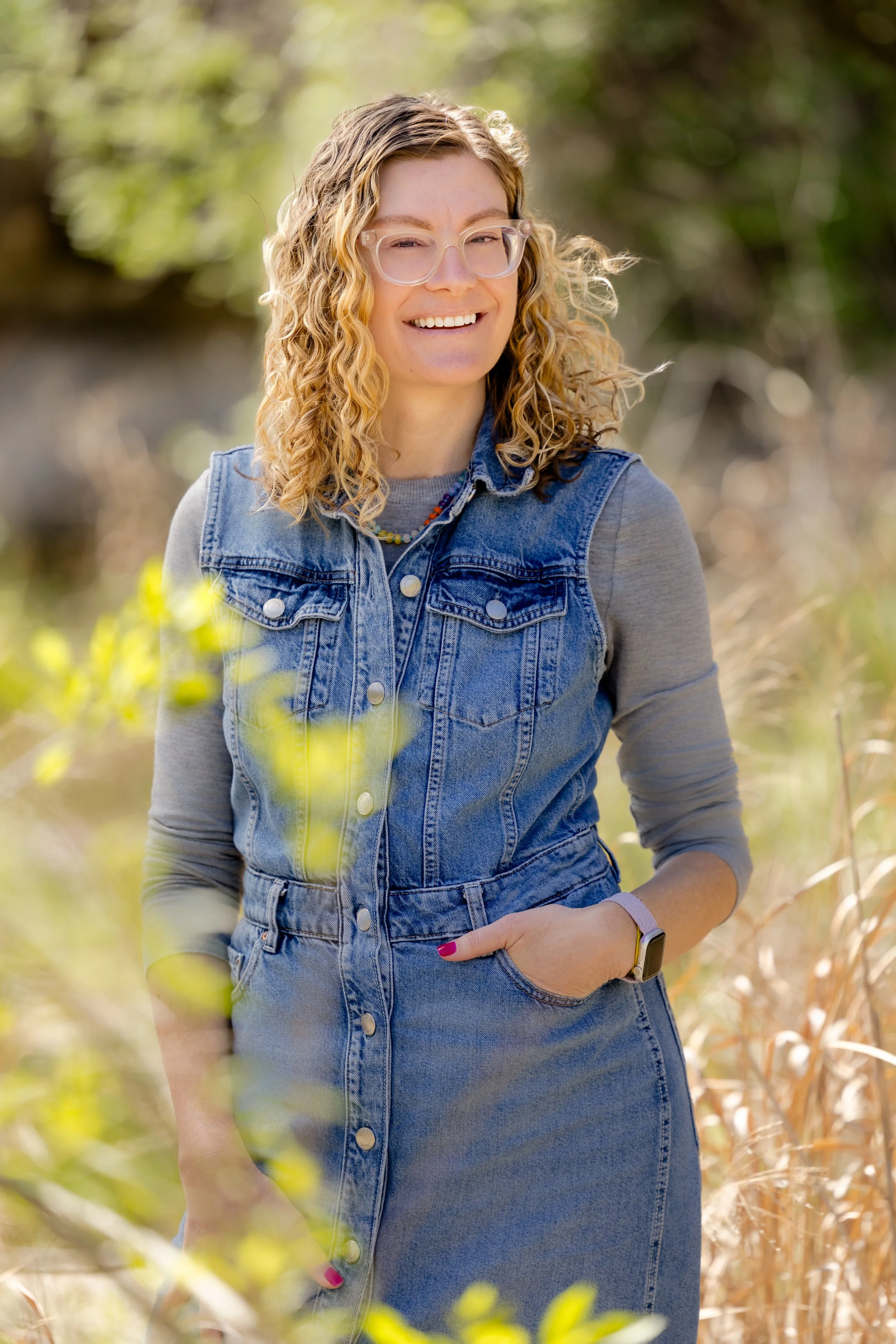 A woman with curly blonde hair, wearing glasses, a denim vest, and a smartwatch, smiling outdoors in a natural setting with blurred trees and plants in the background. Photo by Liza Hippler.