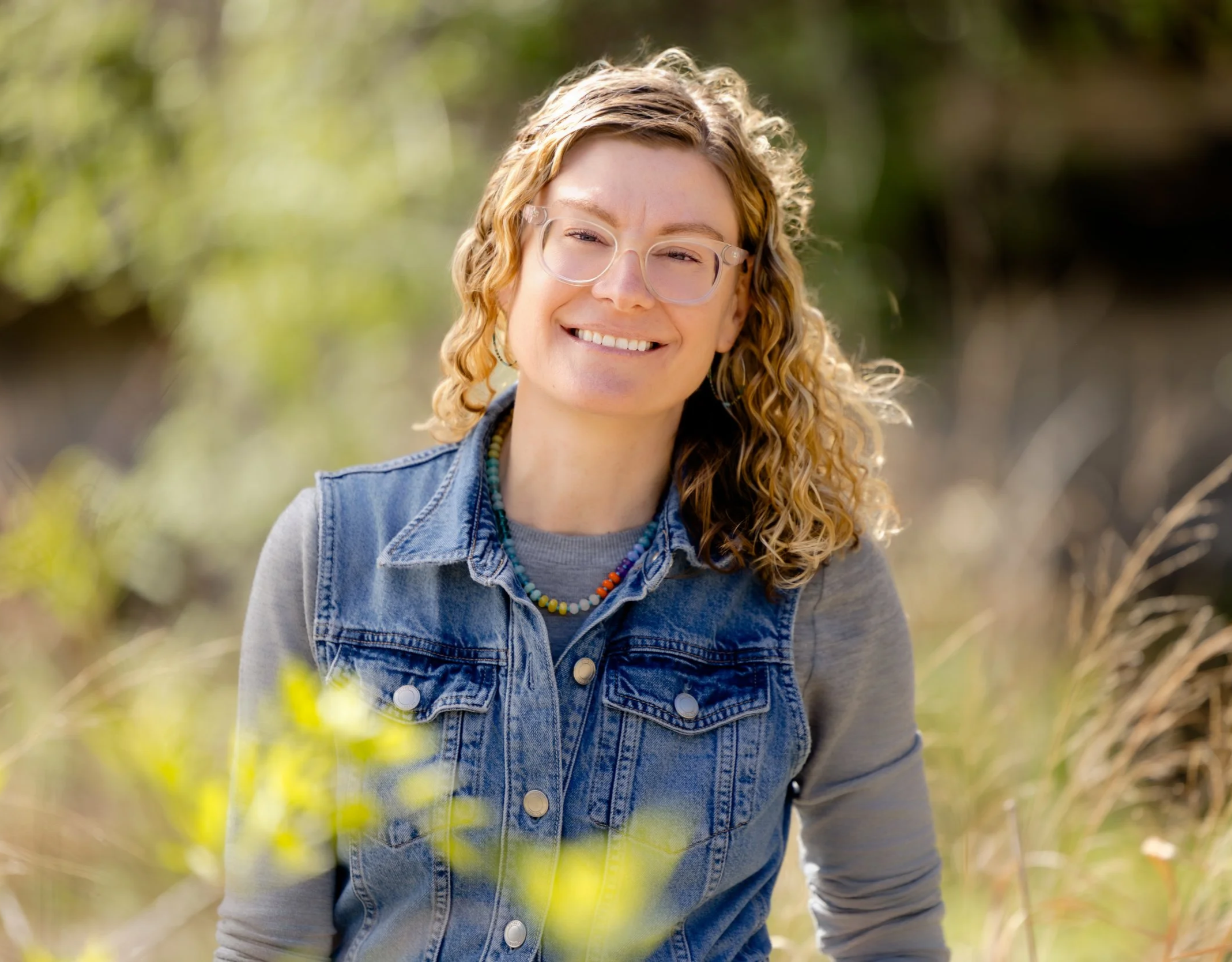 A young woman with curly red hair, wearing glasses, a denim vest, and a colorful beaded necklace, smiling outdoors with blurred greenery in the background.