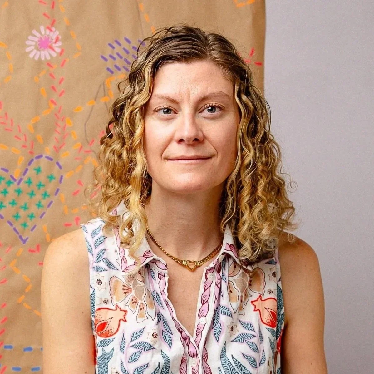 A woman with curly blonde hair wearing a sleeveless floral blouse and a gold heart-shaped necklace, standing in front of a colorful abstract wall art. Picture by Liza Hippler.