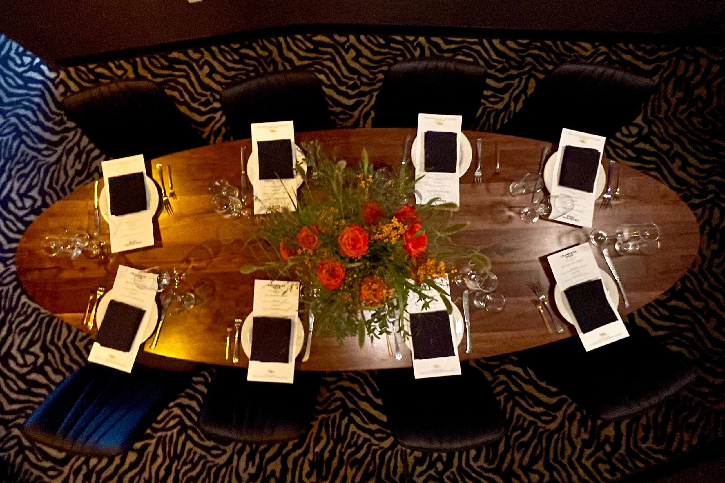 Round banquet table with black leather chairs on a zebra-stripe patterned carpet, set with white plates, black napkins, silverware, wine glasses, and menu cards, with a large floral centerpiece of orange and yellow flowers and green foliage.