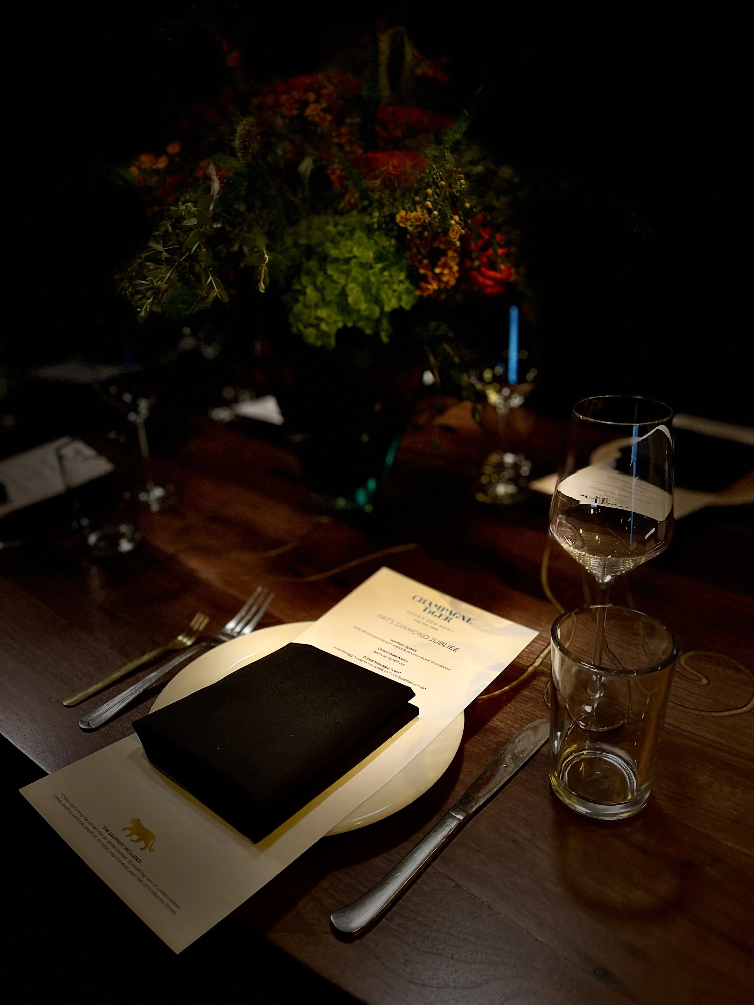 A dimly lit table setting with a flower arrangement in the background, a black gift box on a white plate, a menu, and glasses of water, suggesting a fancy dinner or celebration.