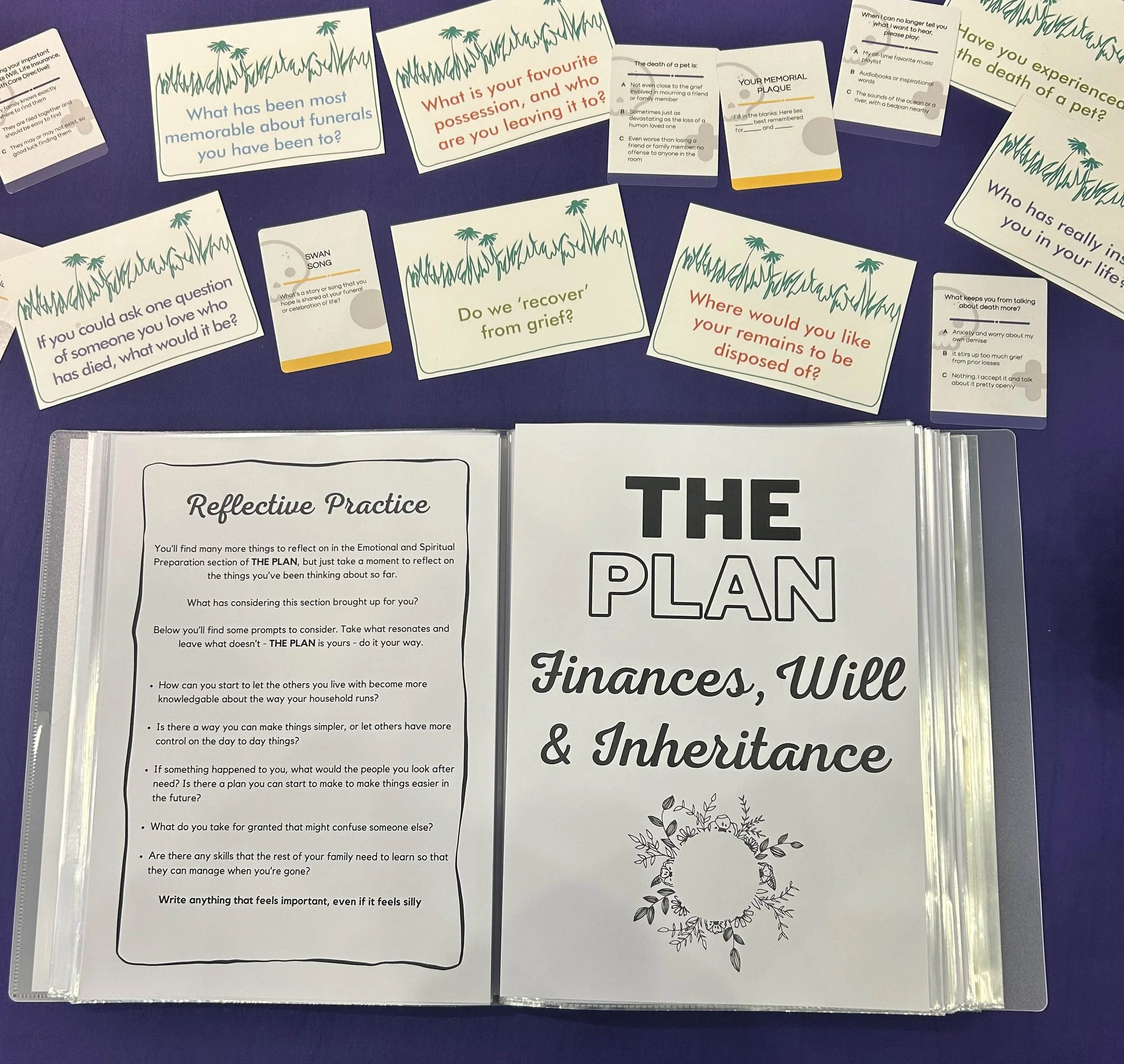 Collection of educational and reflective cards on a blue table, with titles about grief, funerals, memories, and planning, some featuring decorative grass and floral graphics.