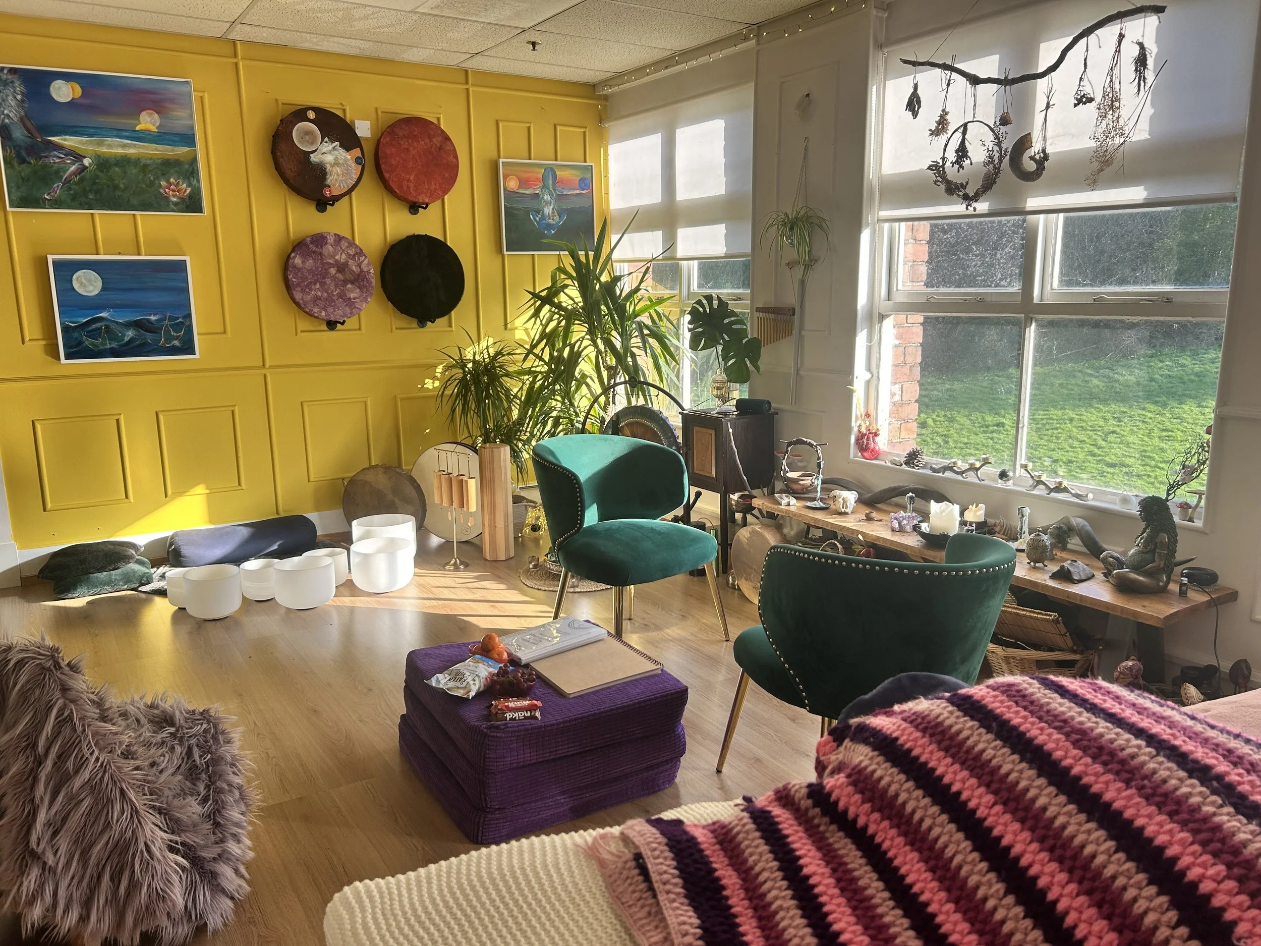 Cozy living room with yellow accent wall displaying colorful paintings, surrounded by plush chairs, potted plants, and decorative items. Large windows let in natural light and offer a view of greenery outside.