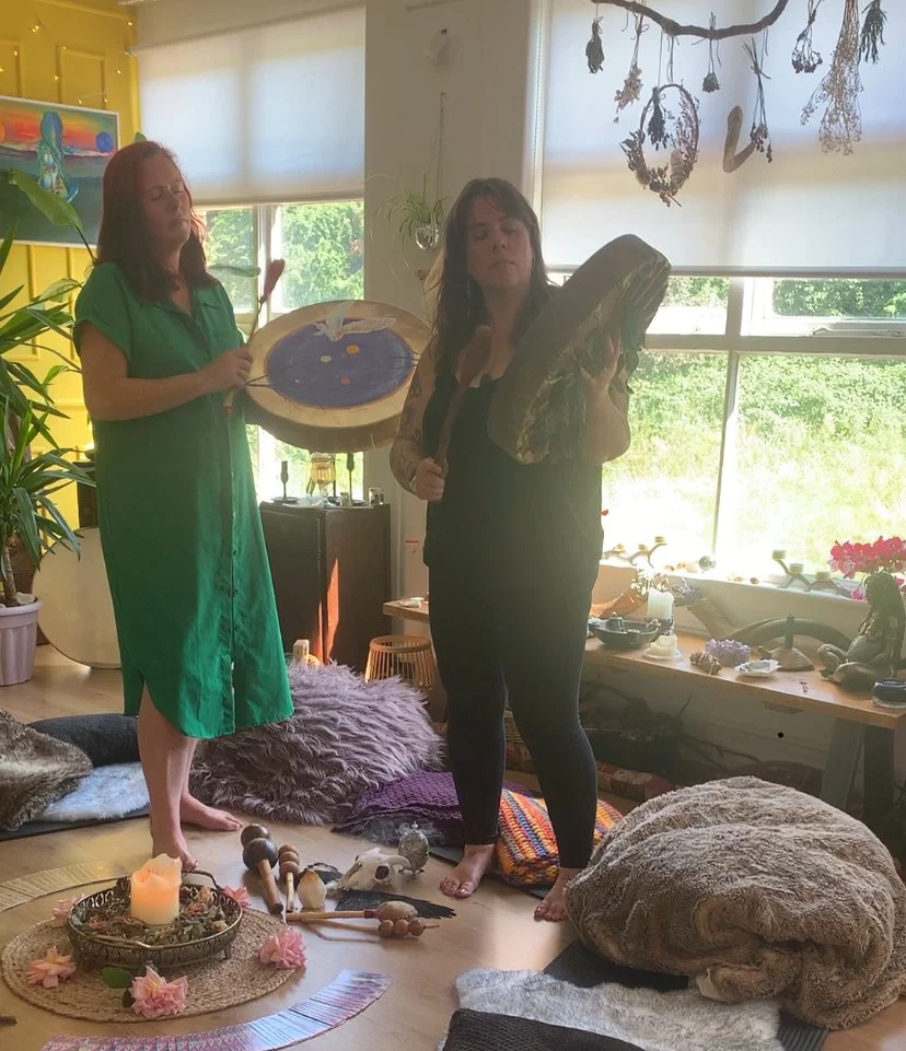 Two women in a cozy, decorated room with spiritual objects and plants. Both women are playing frame drums. The room has natural light, colourful cushions, and various decorative items.