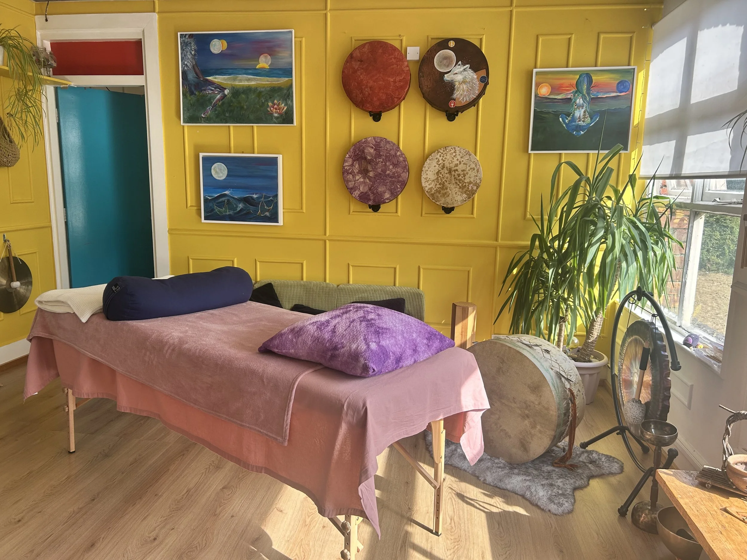 A cozy room with bright yellow walls decorated with colorful paintings, a large window letting in natural light, and various plants and objects including a massage table with a pink sheet, pillows, and drums with distinctive designs.