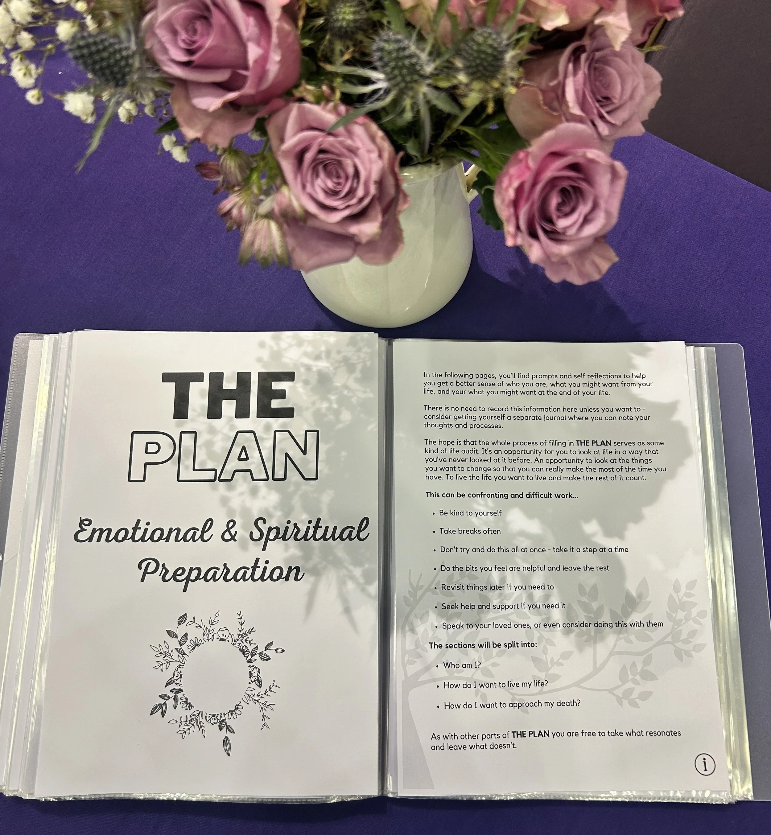 Open book with the title 'The Plan: Emotional & Spiritual Preparation' placed on a purple surface. Above the book is a flower arrangement in a white vase, containing pink roses, small white flowers, and green thistles.