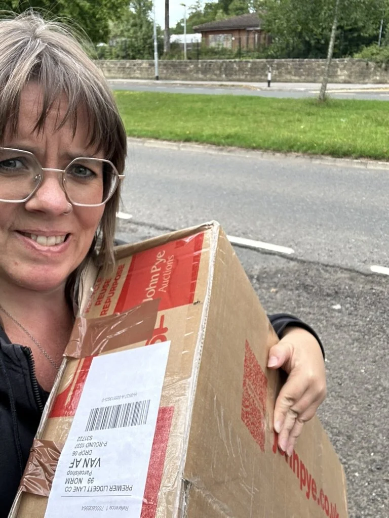 Eva is a white middle aged woman with increasingly spangly hair, carrying a large box under her arm. She looks slightly concerned about how she’s going to carry the box home.