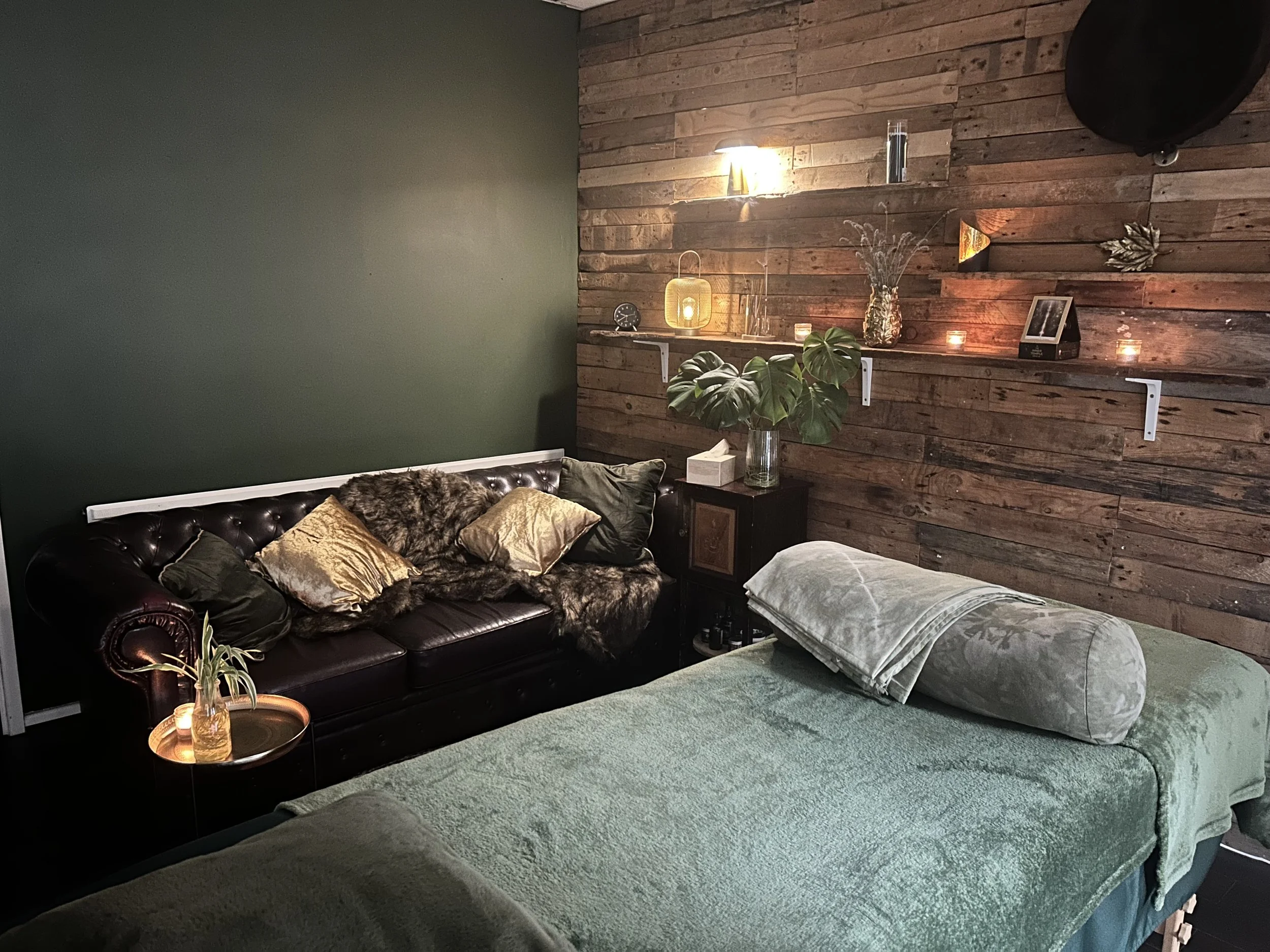 A cozy living room with a dark leather tufted couch adorned with gold and dark pillows and a rustic wooden accent wall decorated with candles, lamps, a clock, plants, and decorative items.