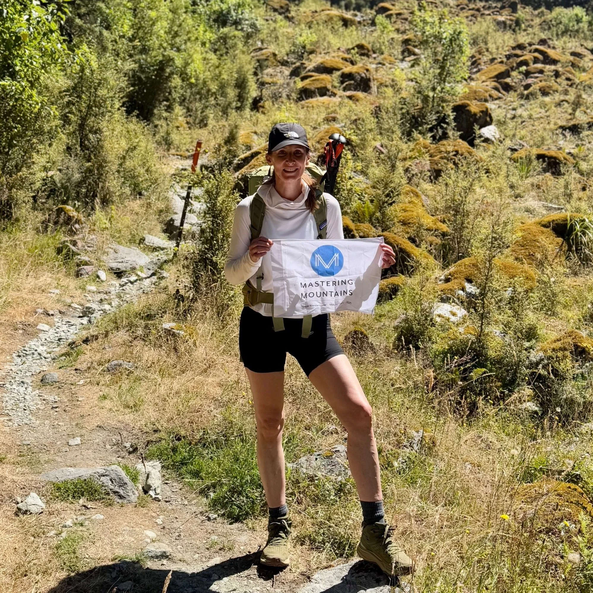 A woman standing on a mountain trail holding a 'Mastering Mountains' banner, wearing hiking gear and a backpack, with greenery and rocks in the background.