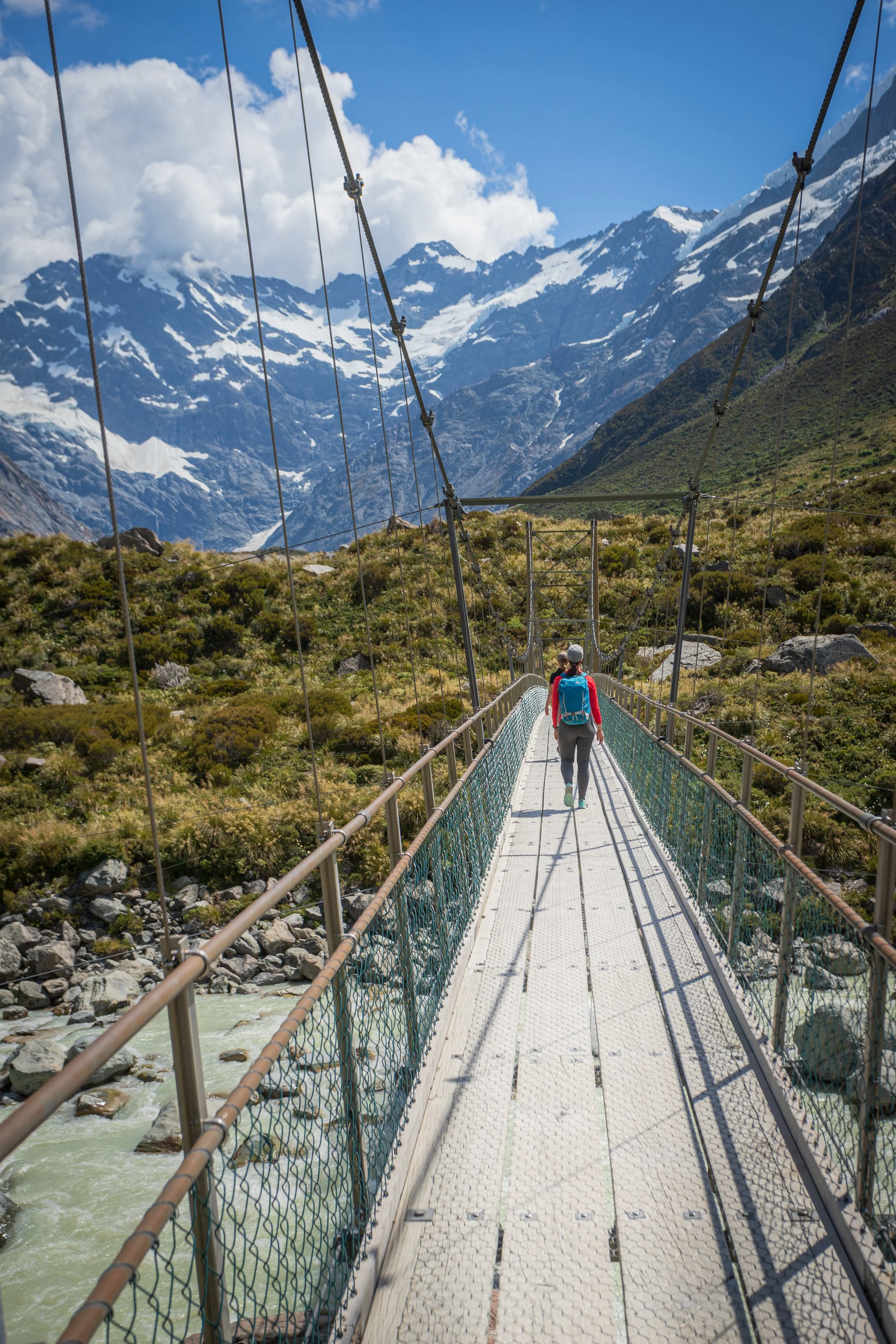Hiker walking on a suspension bridge over a river, with snow-capped mountains and cloudy sky in the background.