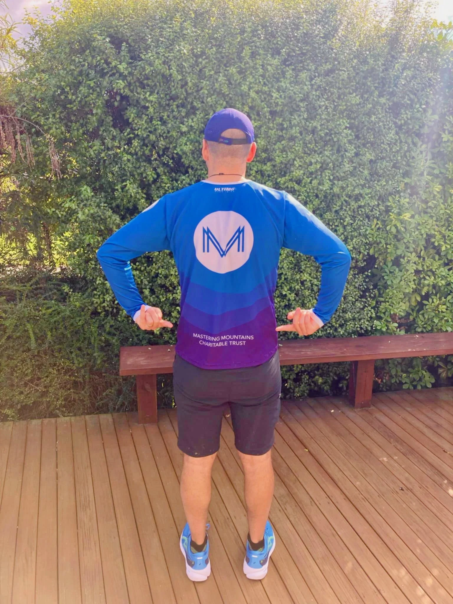 Man wearing a blue long-sleeve shirt, black shorts, a blue cap, and running shoes, standing on a wooden deck with his back to the camera. He is flexing his arms and pointing to the logo on his shirt, with green bushes in the background.