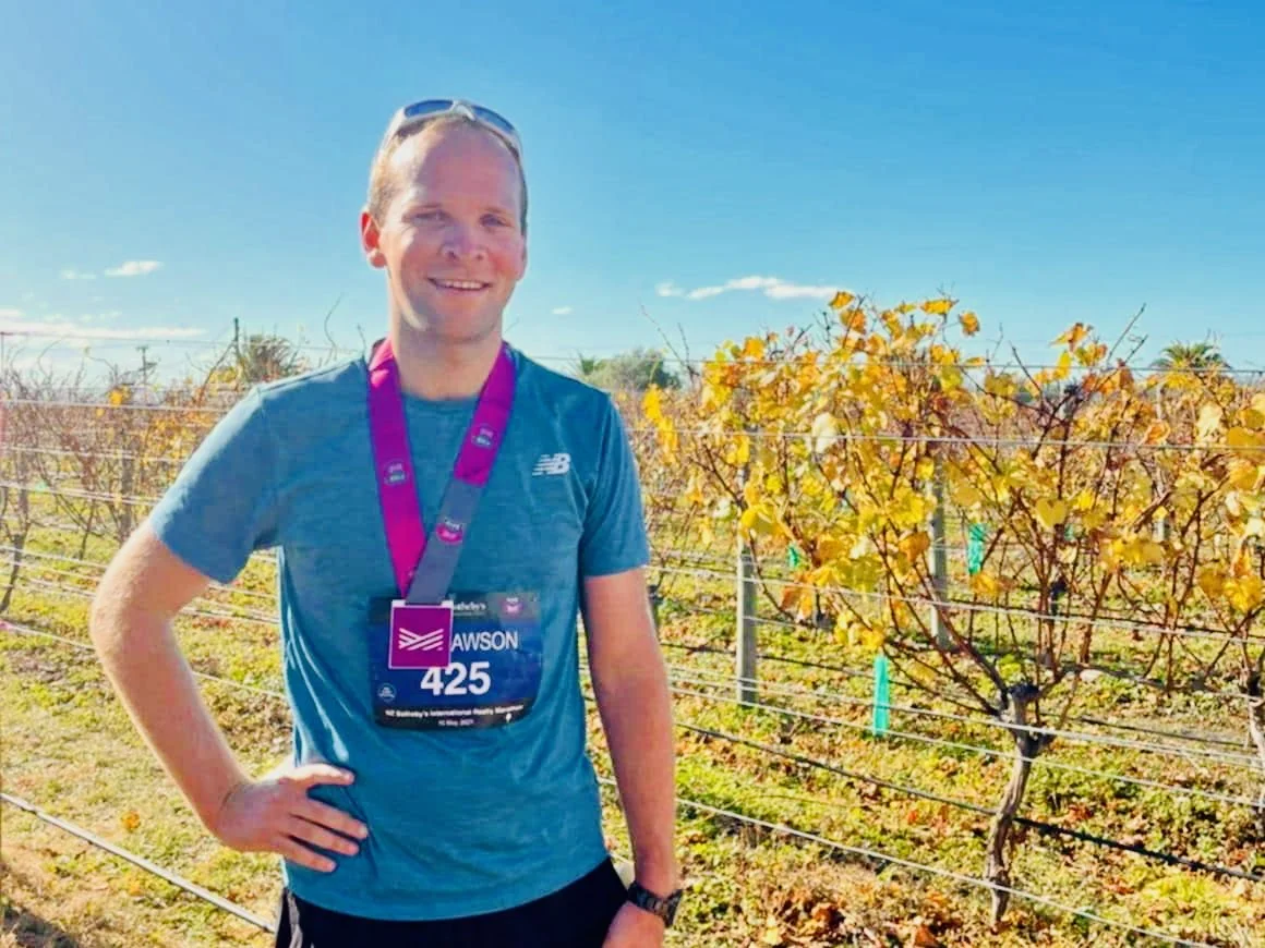 A man with a race bib stands outdoors in a vineyard with yellowing grapevines, smiling at the camera on a sunny day.