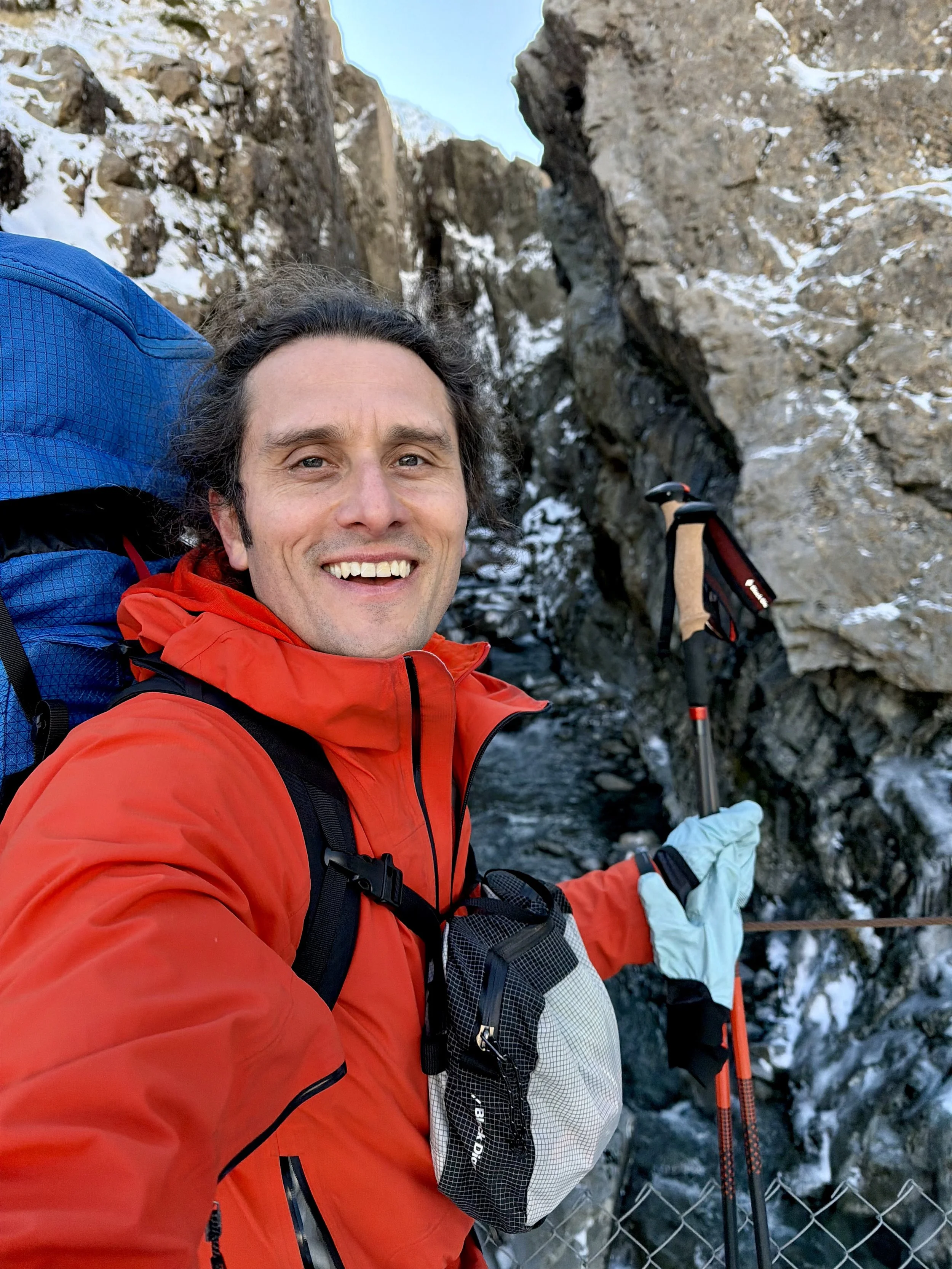 A smiling man in a green athletic jacket taking a selfie outdoors with mountains, trees, and a lake in the background.