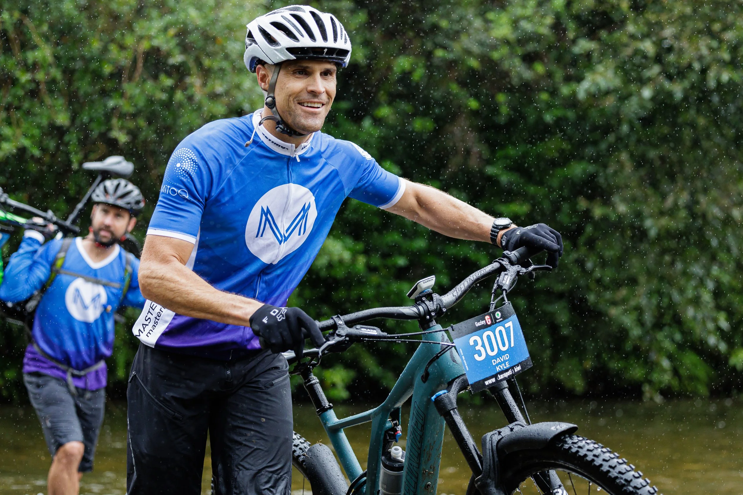 David Kyle and a friend, wearing blue cycling jerseys and helmets on mountain bikes, with one in the foreground smiling and pushing his bike through a forested area while rain falls.