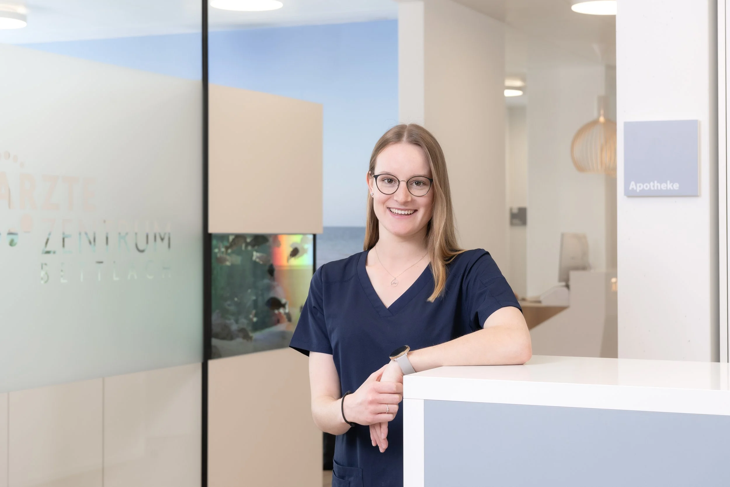 Porträt einer lächelnden Frau mit Brille und langen blonden Haaren vor blauem Hintergrund.