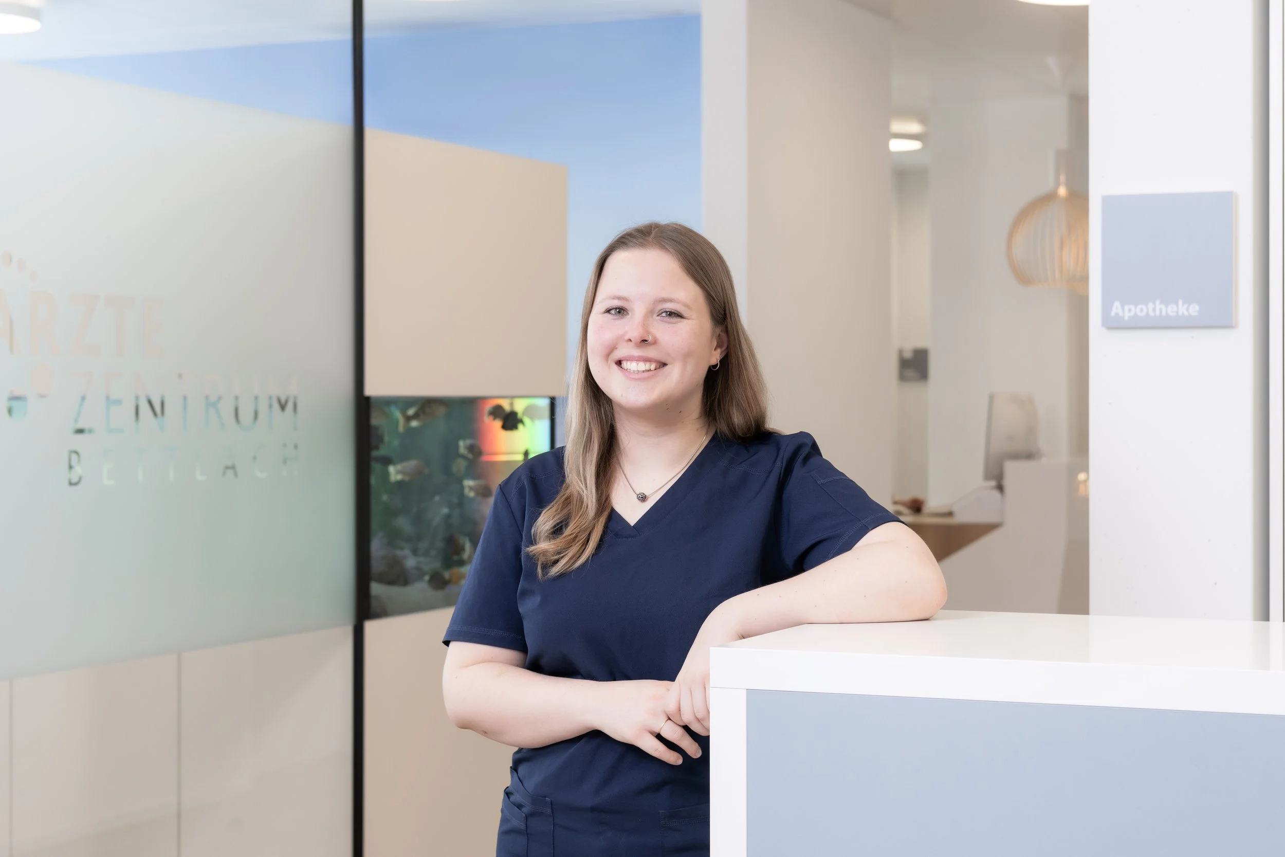Porträt einer lächelnden Person mit langen blonden Haaren und Nasenpiercing vor einem blauen Hintergrund.