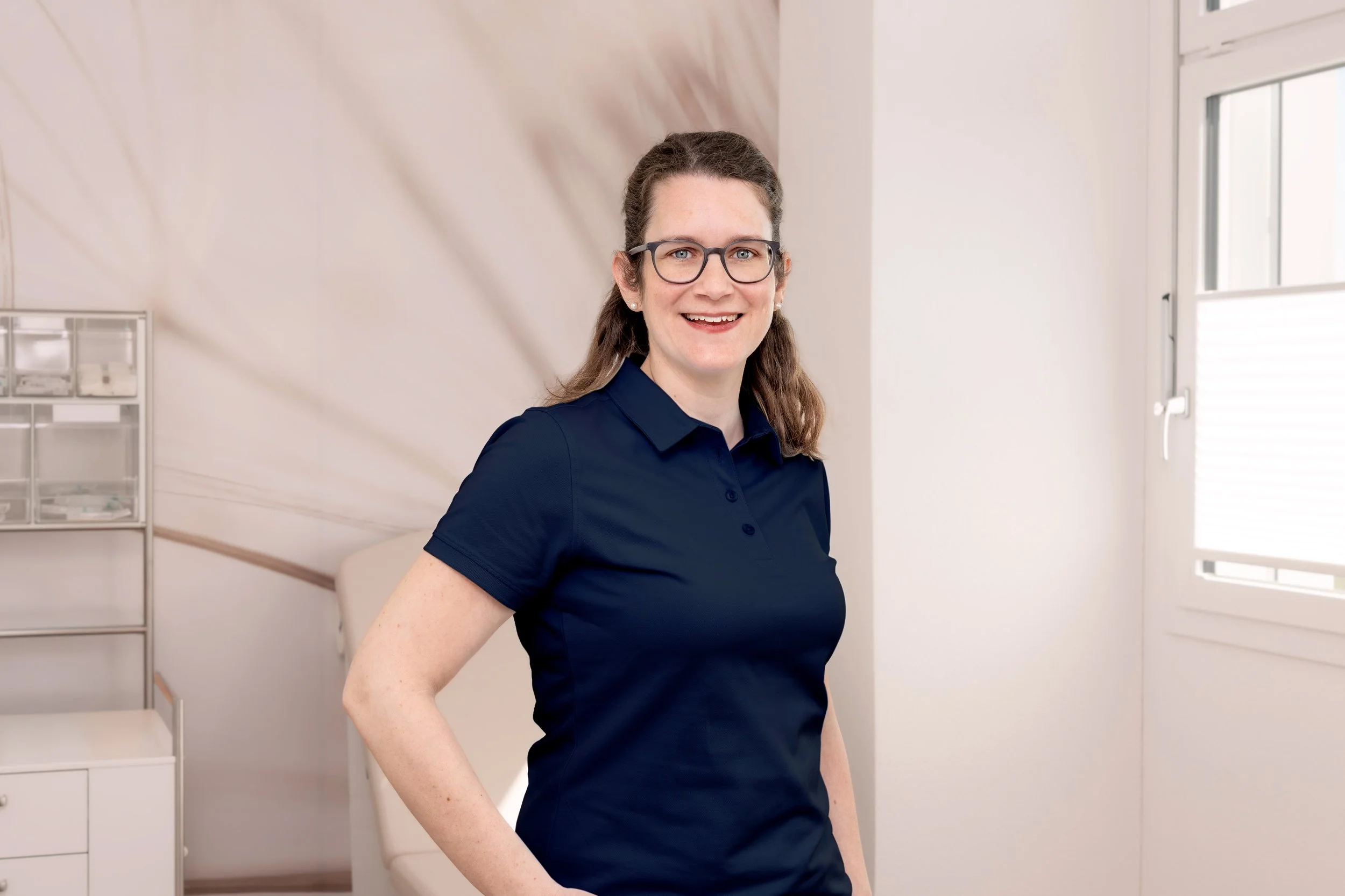 Porträt einer lächelnden Frau mit Brille und langen Haaren, die dunkelblaues Oberteil trägt, vor blauem Hintergrund.