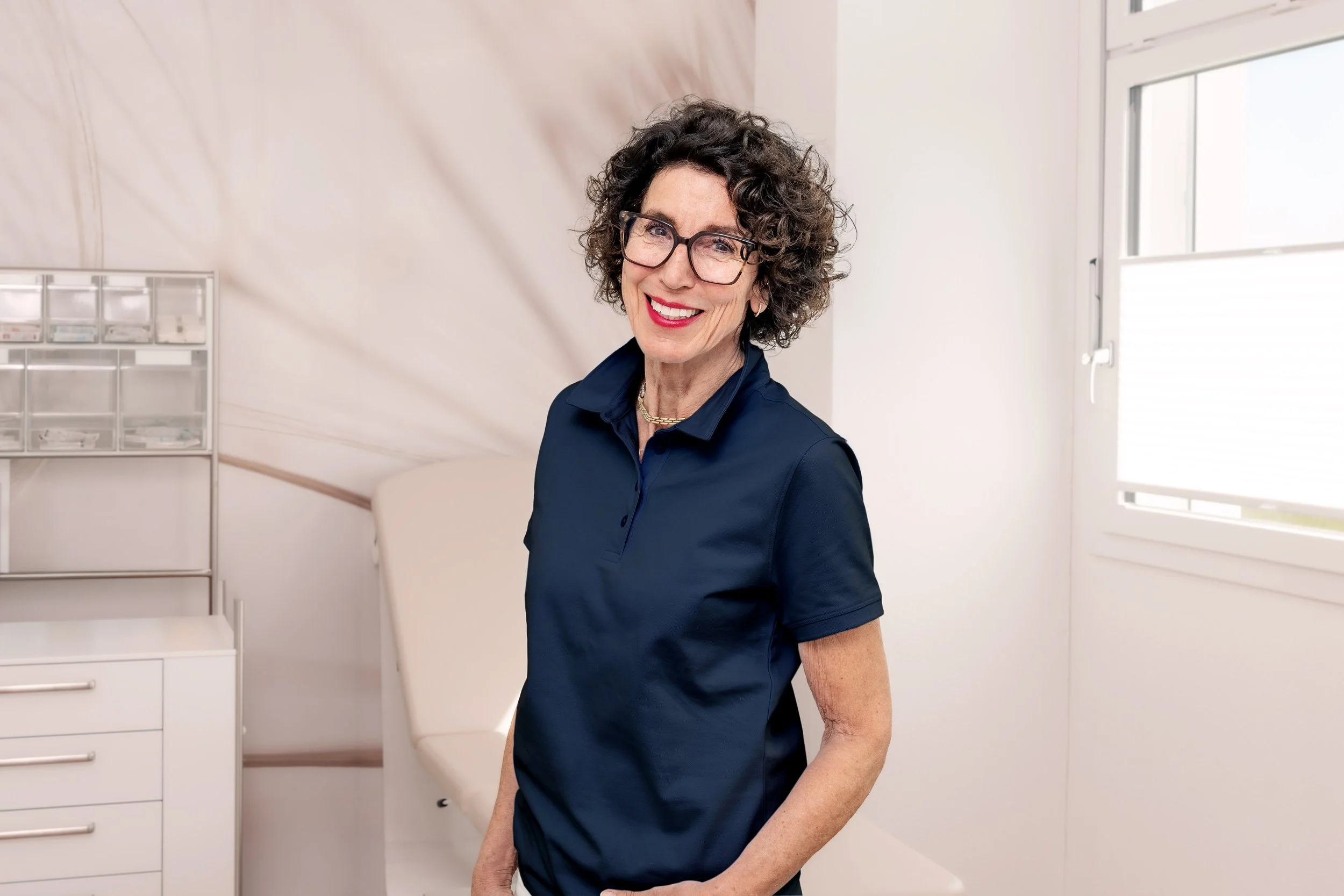 Portrait einer lächelnden älteren Frau mit lockigem Haar und Brille, die einen pinken Pullover trägt, vor einem blauen Hintergrund.