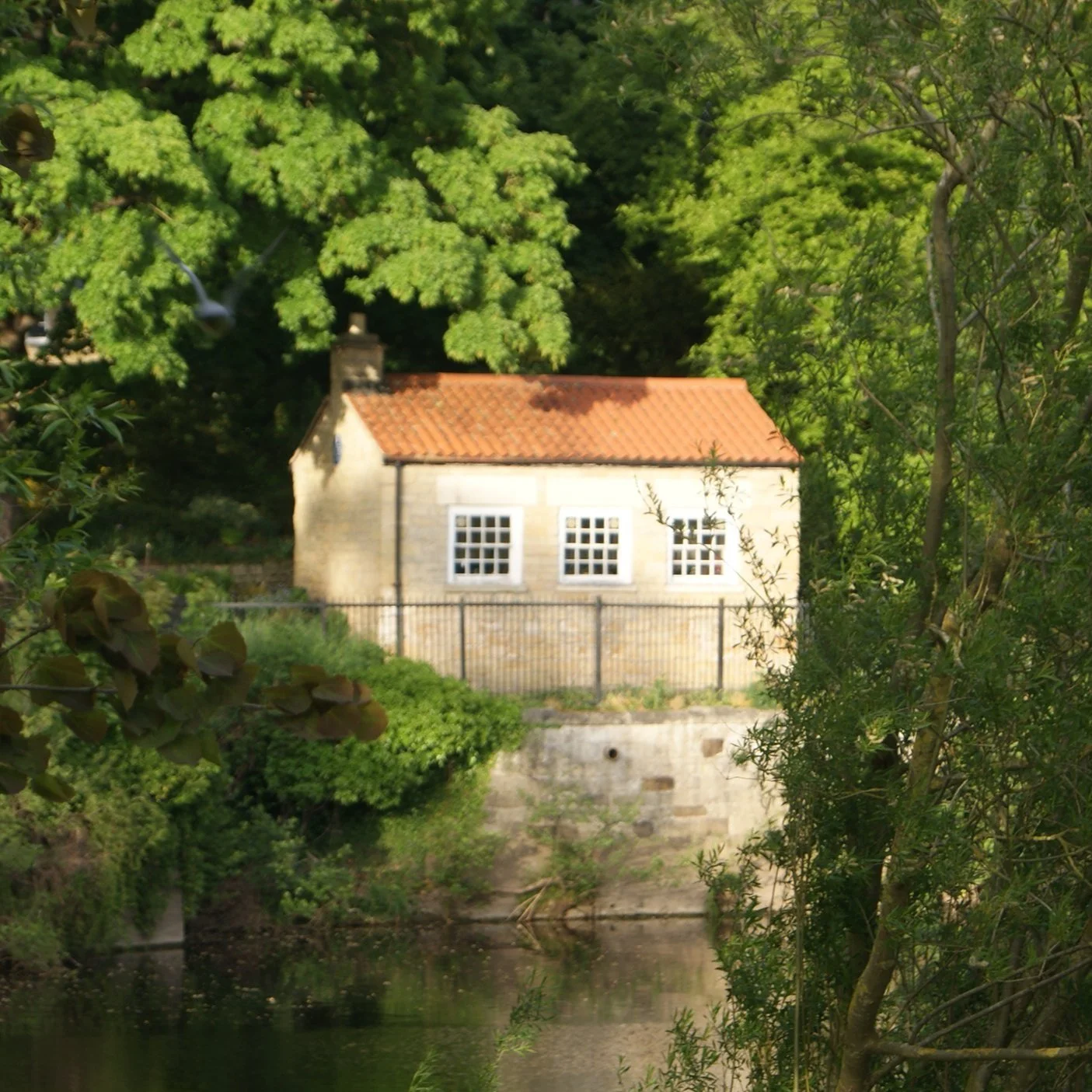 Small stone building with red-tiled roof near a river, surrounded by lush greenery and trees.