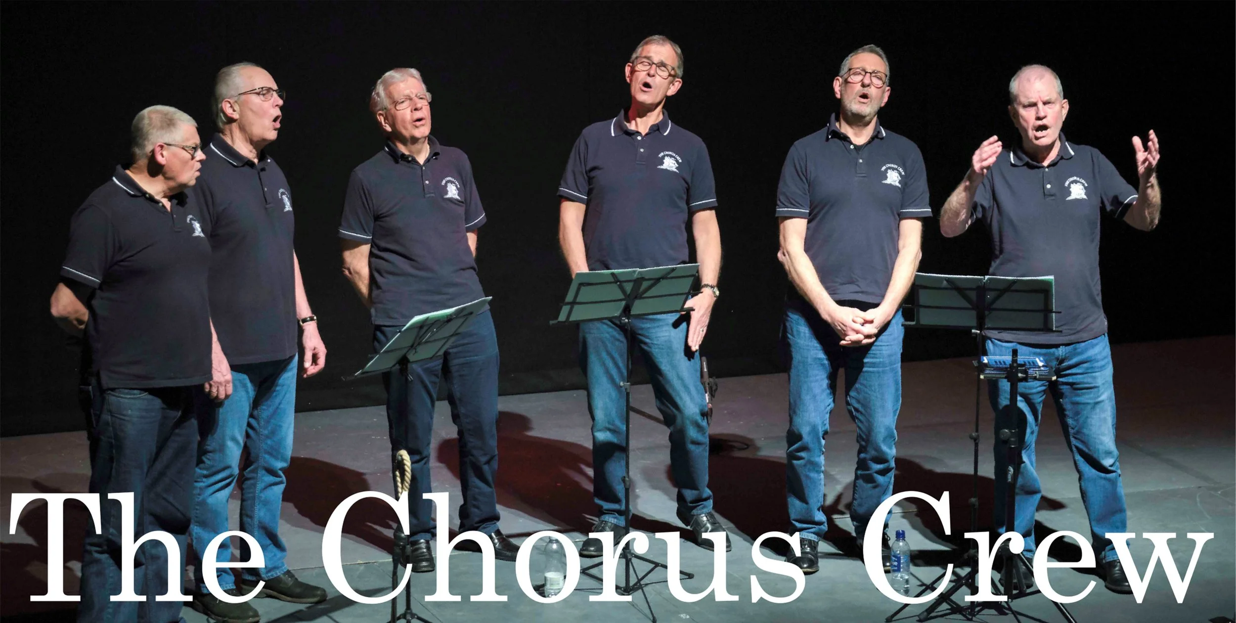 Six men in matching polo shirts and jeans singing on stage, with music stands in front of them. Text reads "The Chorus Crew."