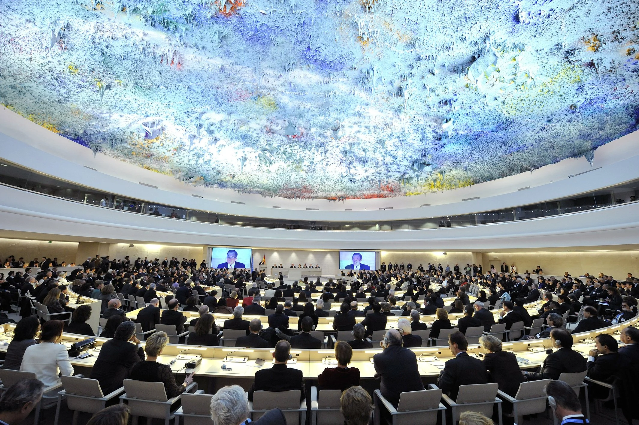 A large conference room filled with numerous people seated at curved desks during a formal meeting or conference. The room has a high ceiling with an elaborate, colorful artistic installation resembling a sky or cloud pattern. There are large screens at the front displaying a speaker, and a stage area with multiple individuals seated behind a long desk.