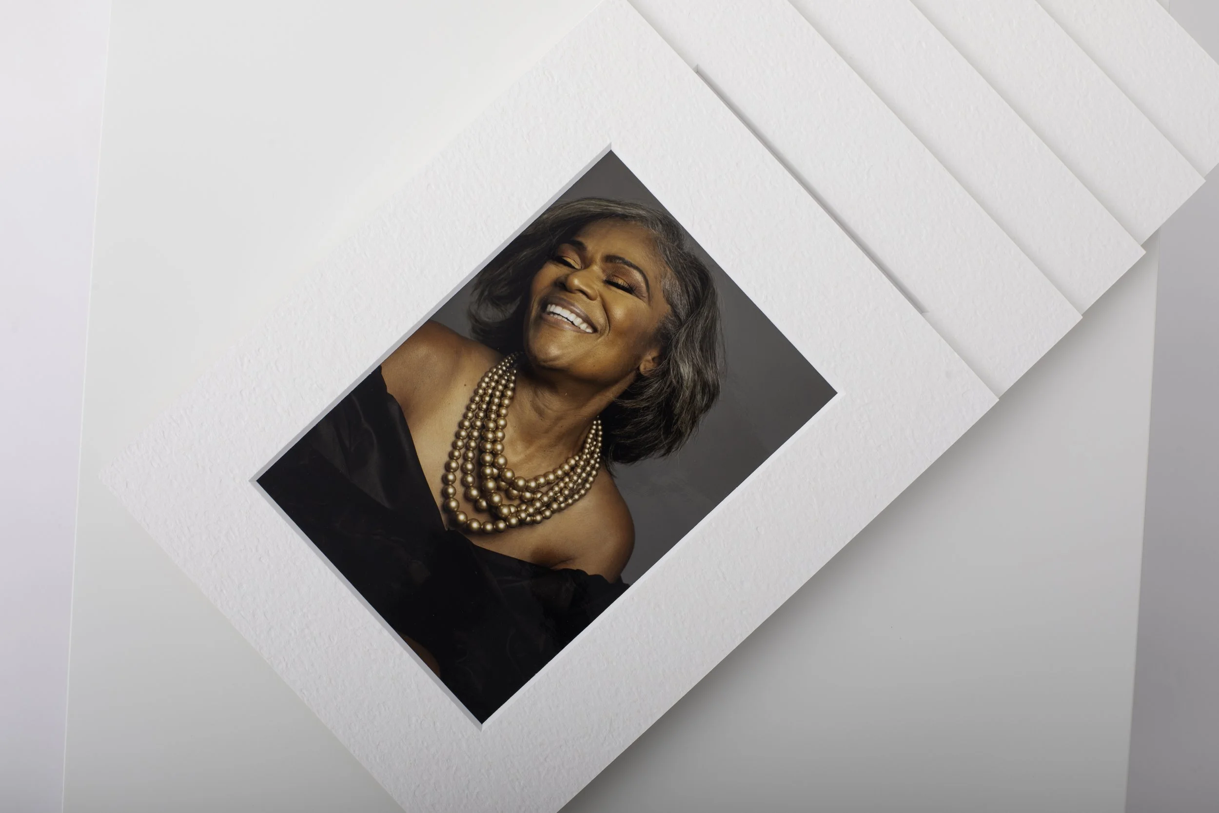 A smiling older woman with gray hair, wearing multiple pearl necklaces and a black off-shoulder top, posing against a gray background.