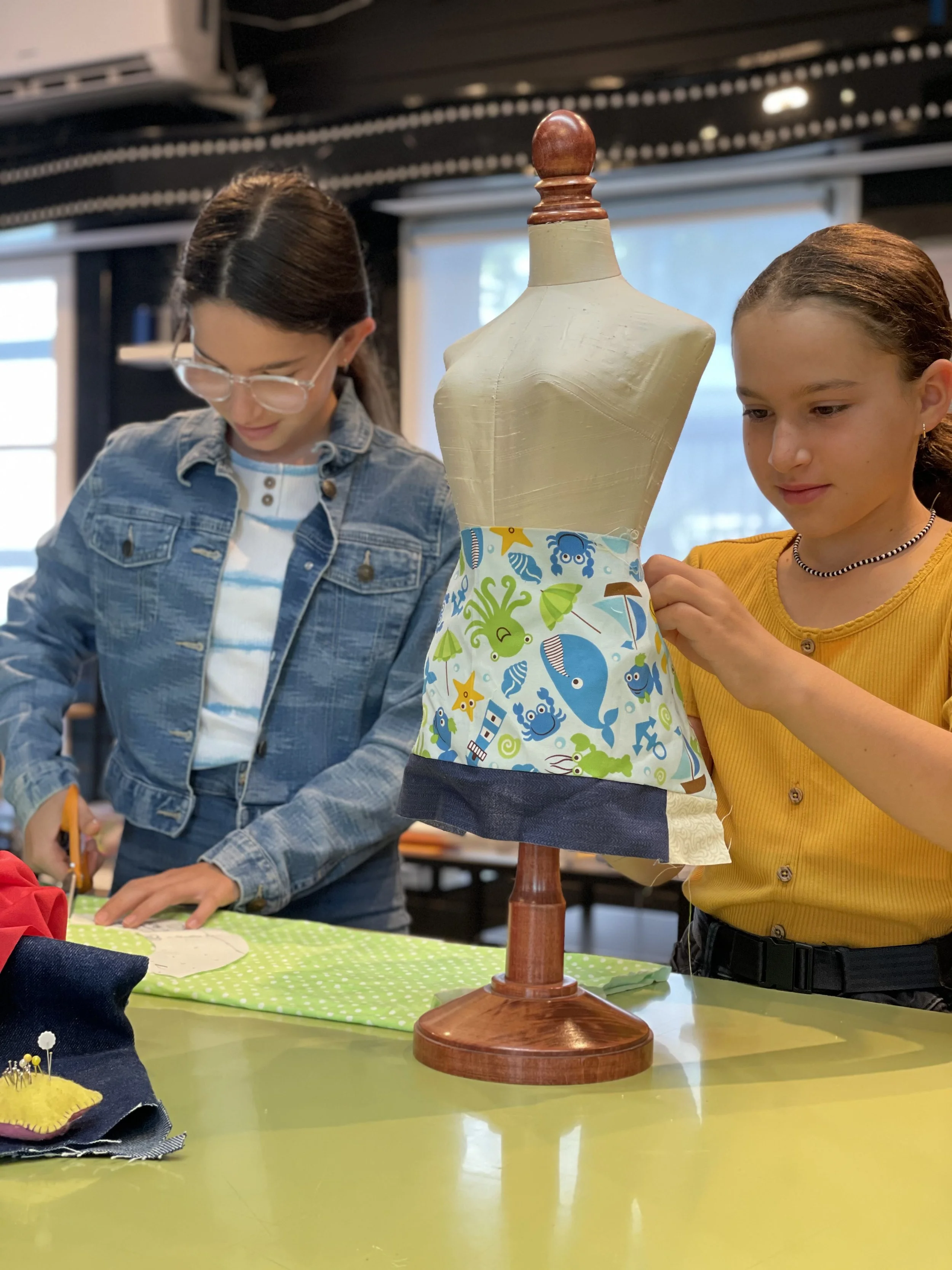 Two young girls working on a fabric project at a table in a room with natural light. One girl is cutting fabric, and the other is adjusting a fabric skirt on a dress form that displays a colorful, ocean-themed fabric.