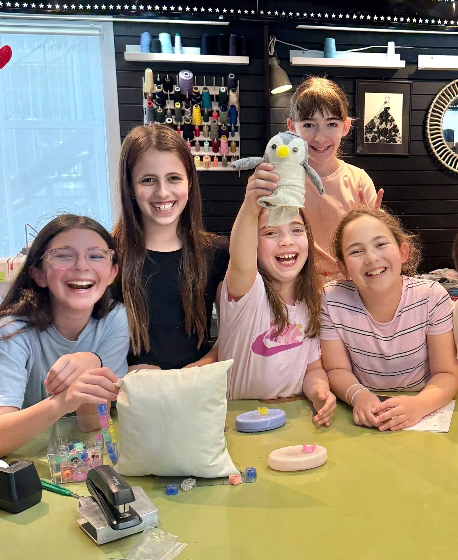 Group of five happy girls making crafts at a table, with one girl holding a sock penguin puppet, in a craft room with sewing supplies and colorful thread spools in the background.