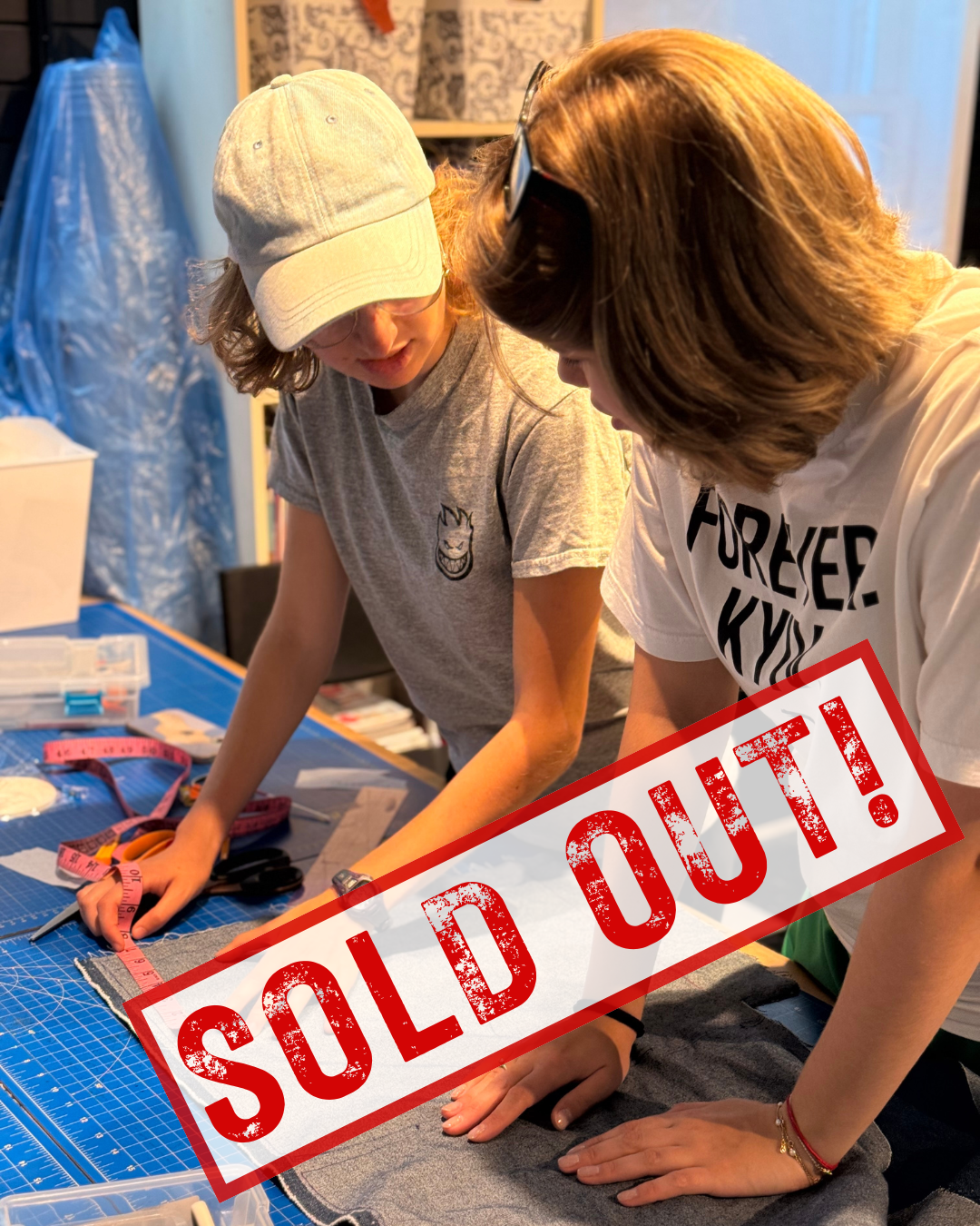 Two women are working together on a sewing project at a craft table, with sewing tools and fabric, in an indoor setting. There is a red 'SOLD OUT!' overlay on the image.