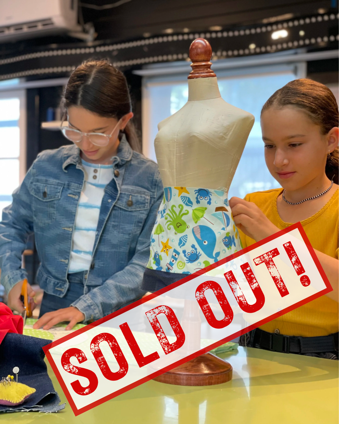 Two young girls working on a craft project with a dress form that has a creative seashell and fish-patterned fabric around its waist. The image has a red "SOLD OUT!" stamp across it.