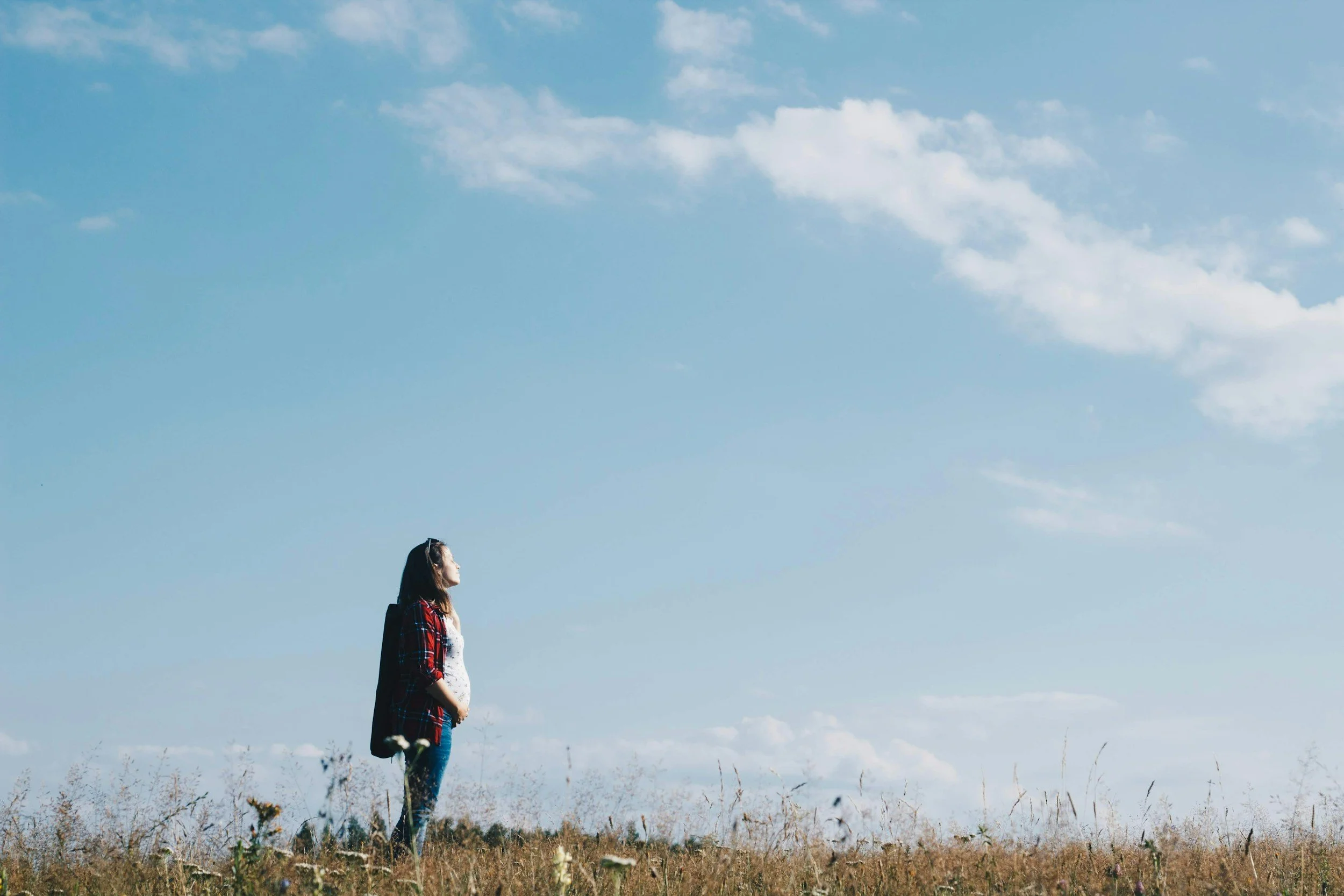 A woman standing alone in a grassy field under a blue sky with scattered clouds.