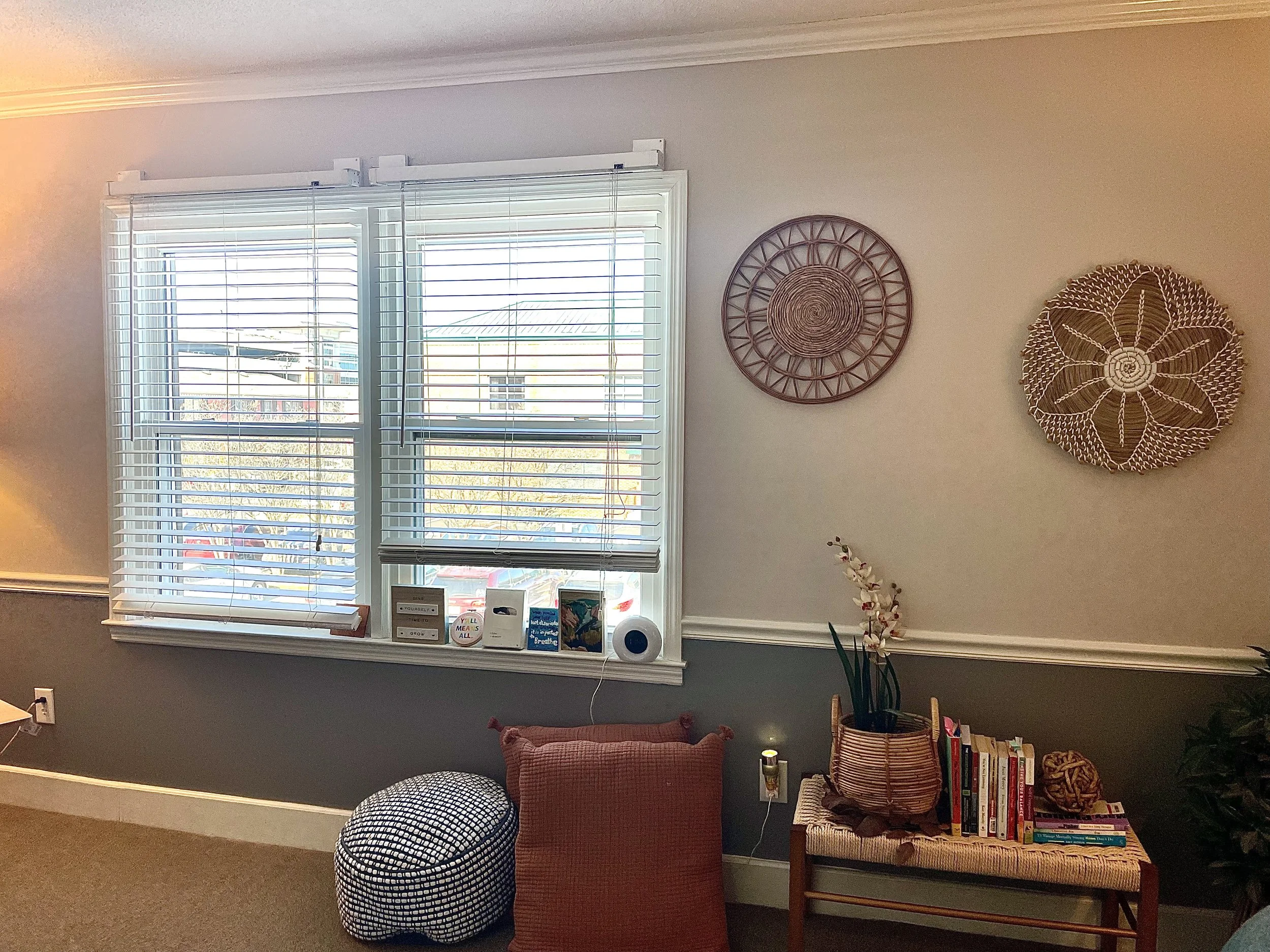 Living room with large window with white blinds, decorated wall art, a small bookshelf with books, a potted plant, and cushions on the floor.